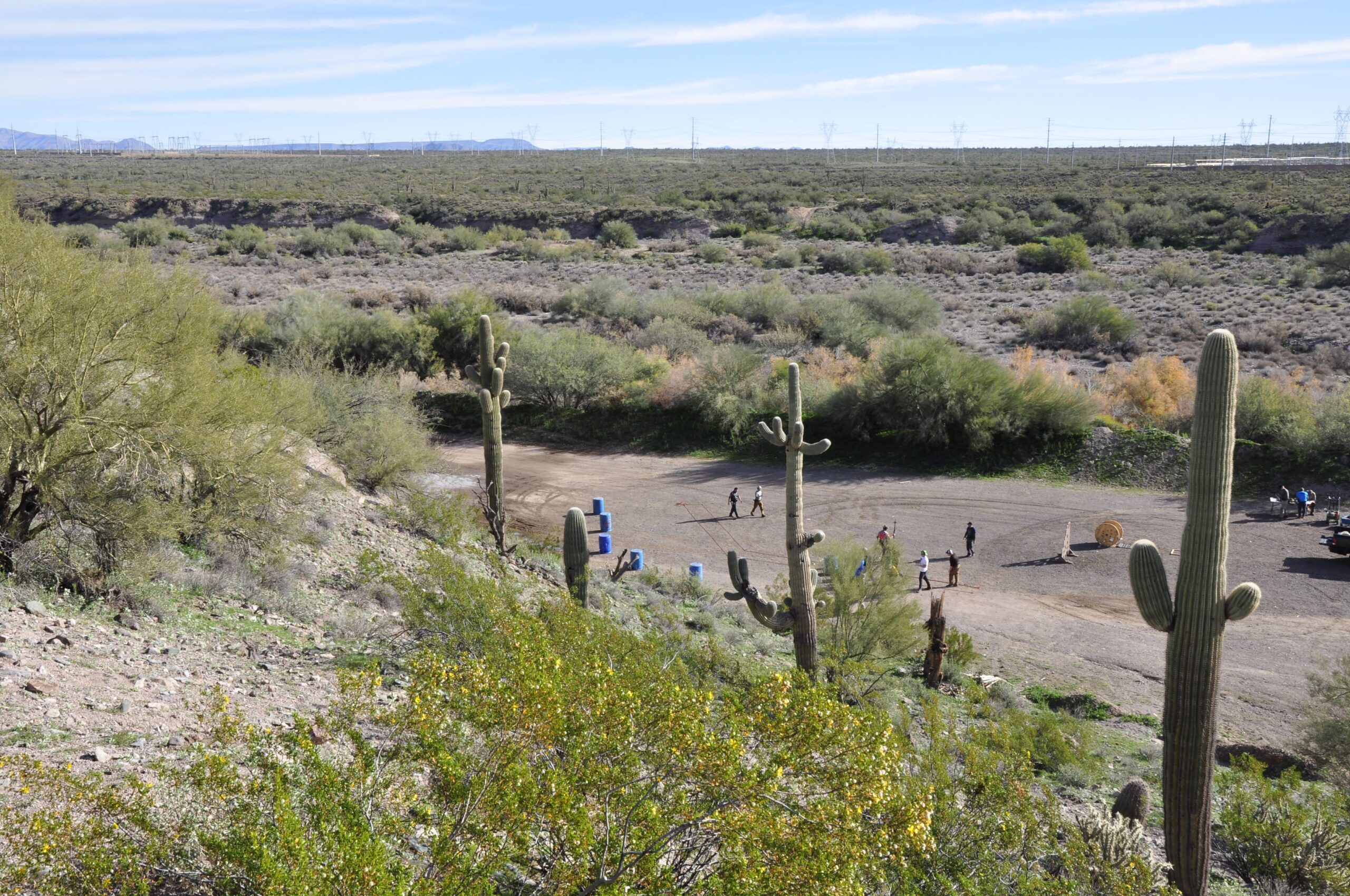 A wide, high-angle shot of a dusty, high-desert outdoor shooting facility. Several shooters are positioned in a large dirt bay equipped with blue barrels, wooden spools, and target stands, all set against a vast landscape of desert scrub and saguaro cacti.