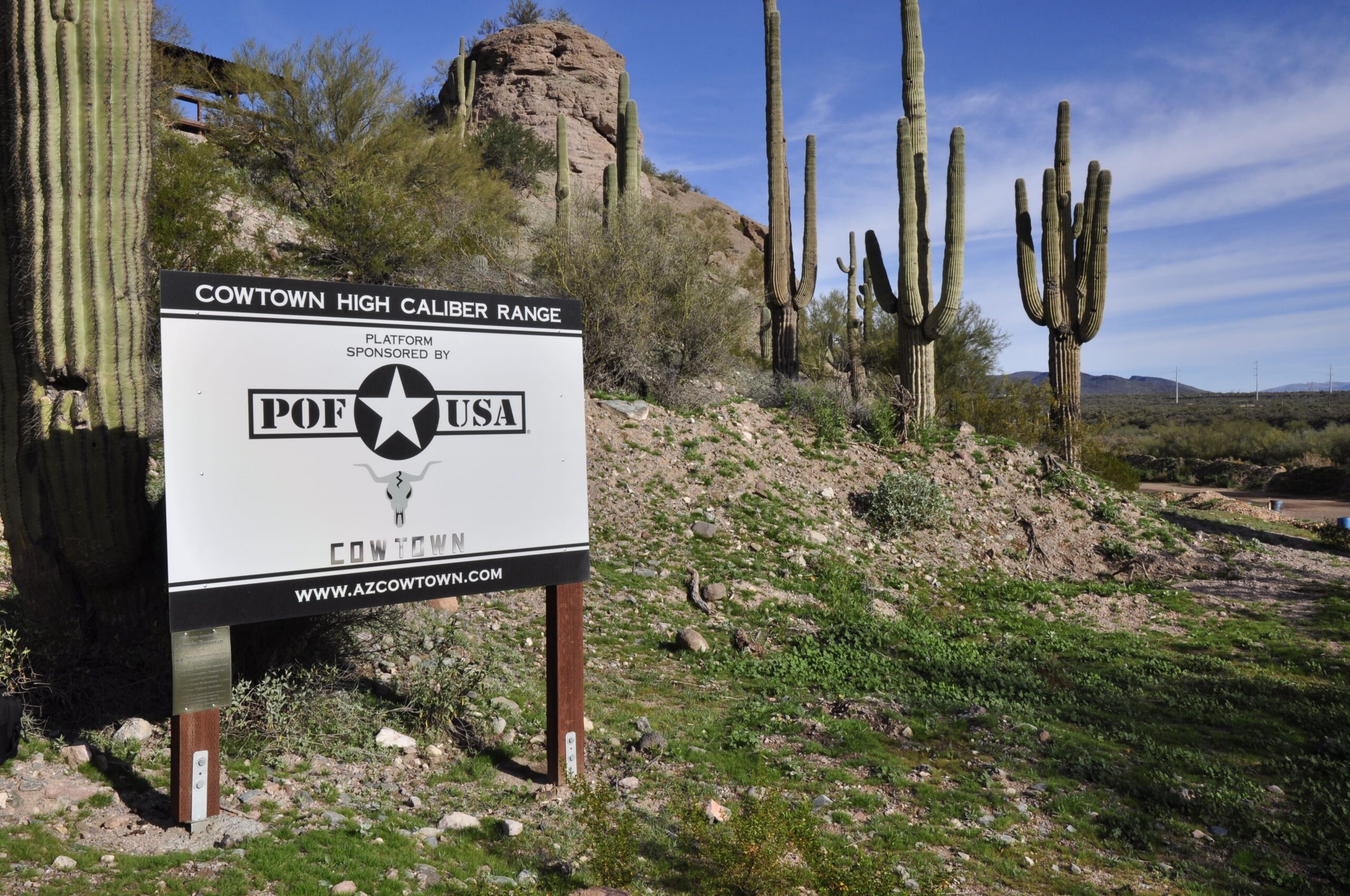 47bb1254-f2e8-4b32-9249-597b5894a0a2.webp A rugged outdoor sign at the high-desert facility identifies the "COWTOWN HIGH CALIBER RANGE" against a backdrop of Saguaro cacti and rocky Arizona terrain.