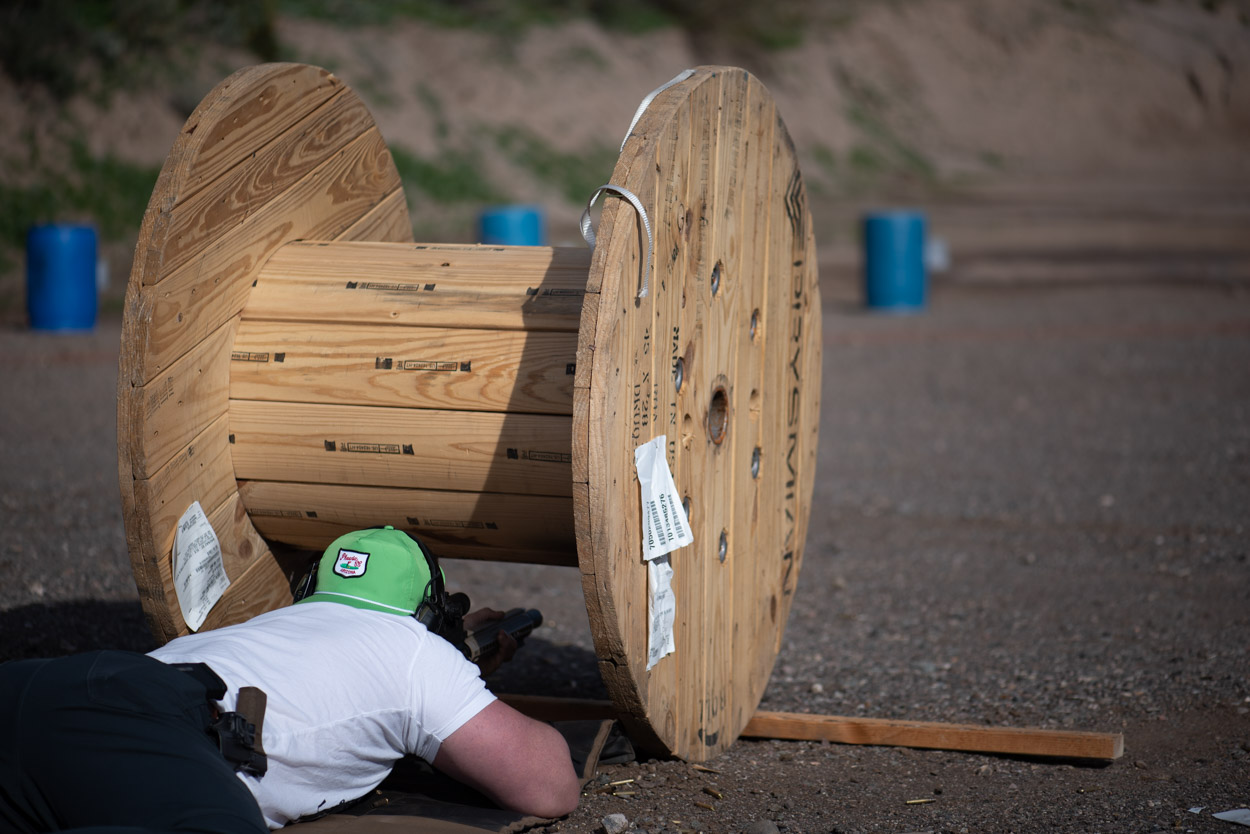 A shooter wearing a green hat lies in a prone position on the dirt, aiming a rifle system through the center of a large wooden cable spool used as a tactical barricade. Scattered brass casings are visible on the ground around the shooter.