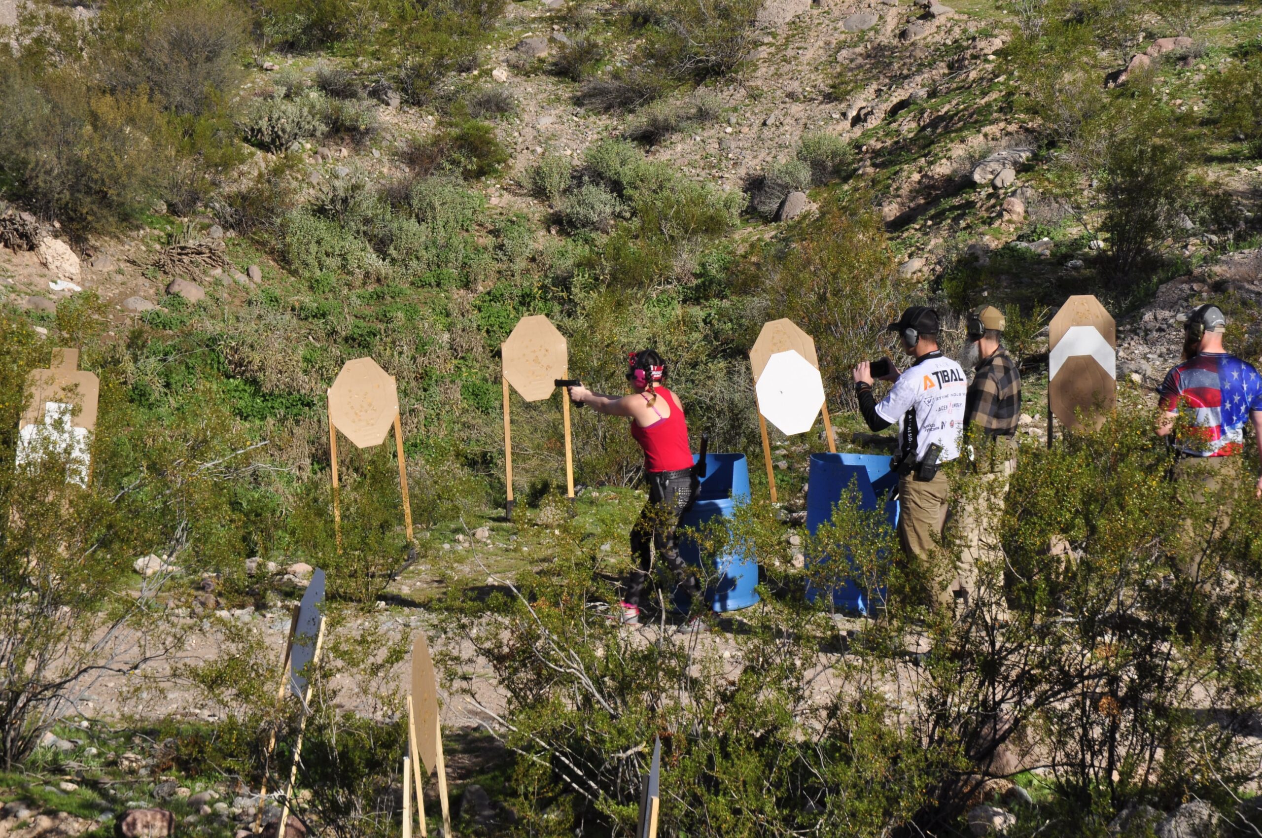 A shooter in motion engages targets in a dusty, high-desert outdoor bay. The image captures a "dirt and brass" aesthetic as multiple members train together in a real-world scenario.