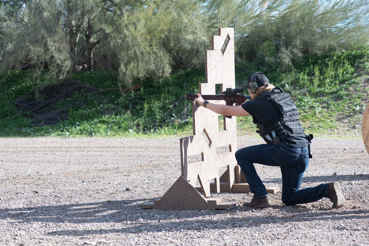 A shooter in a tactical vest and jeans kneels behind a tall, notched wooden barricade, engaging a target with a rifle system. The background shows the dusty ground and green desert vegetation of an outdoor bay.