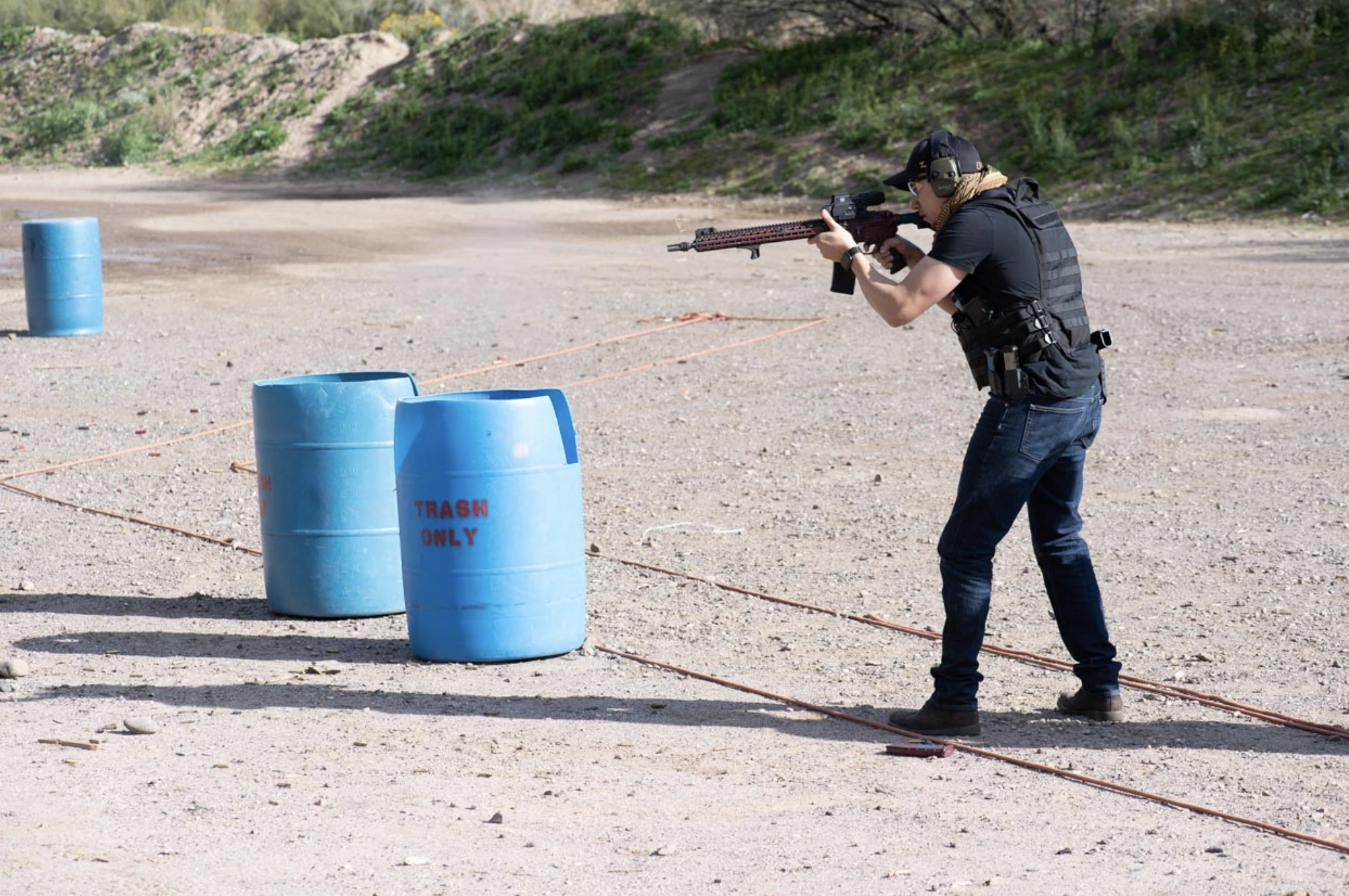 A shooter wearing a tactical vest and hearing protection engaging targets with a rifle platform in a dusty, high-desert bay. The image captures the grit of the Arizona desert environment.