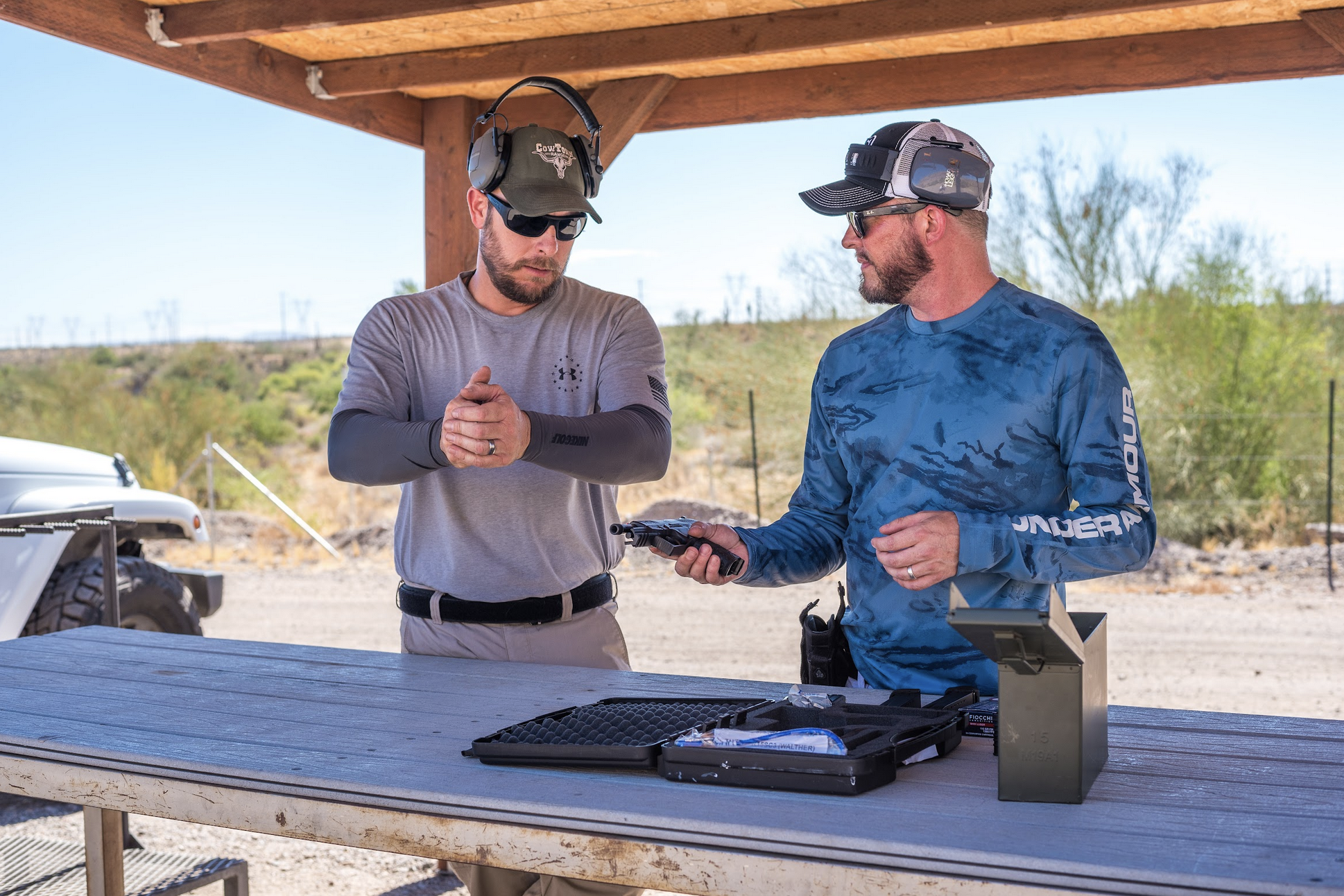 Two shooters stand at a weathered wooden table under a shaded bay, discussing a handgun platform. One shooter, acting as a mentor, demonstrates proper handling techniques to the other.