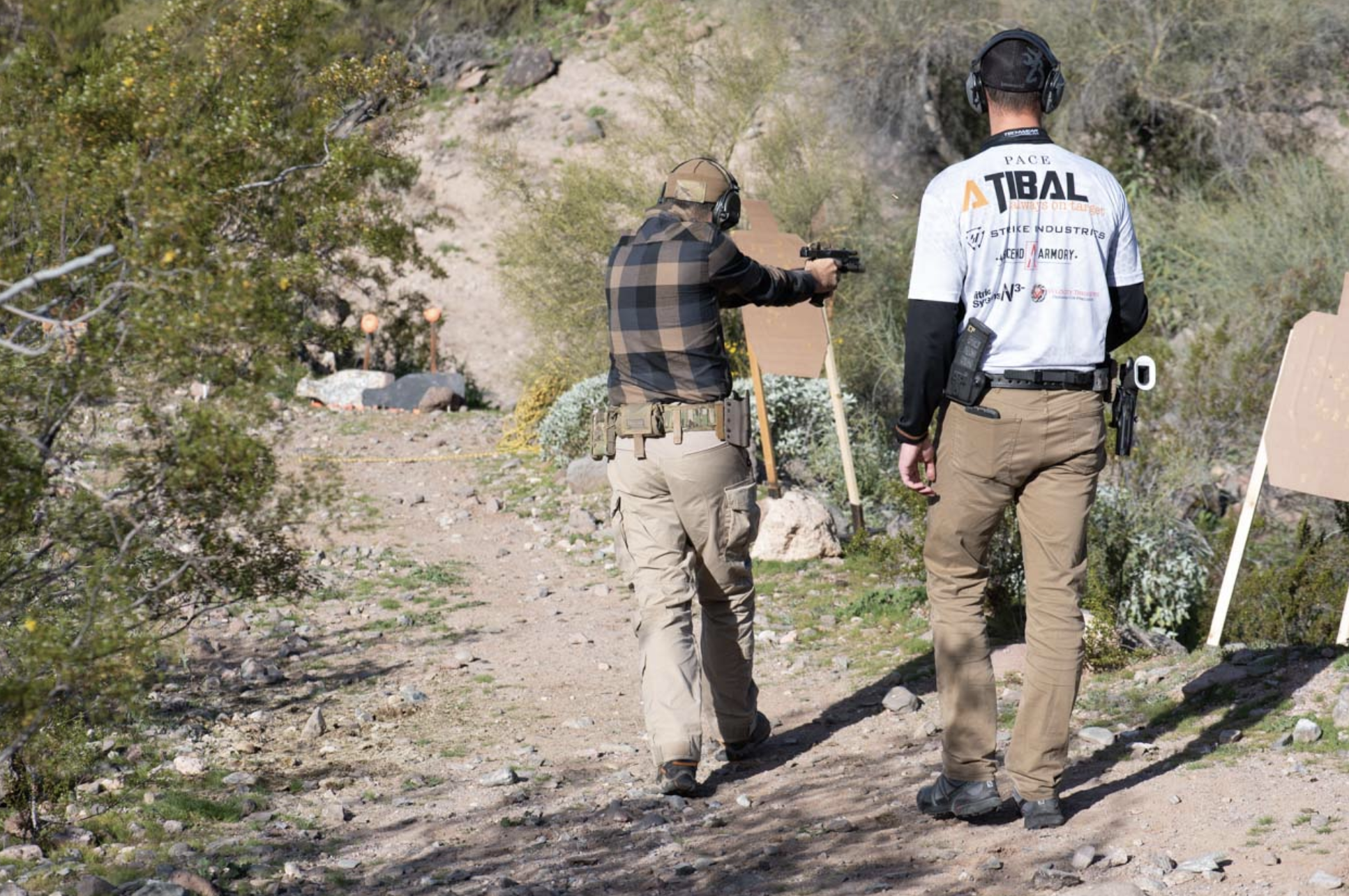 Two men in tactical attire move through a high-desert trail at Cowtown Range. One shooter engages a cardboard silhouette target while the other observes his movement and platform control.