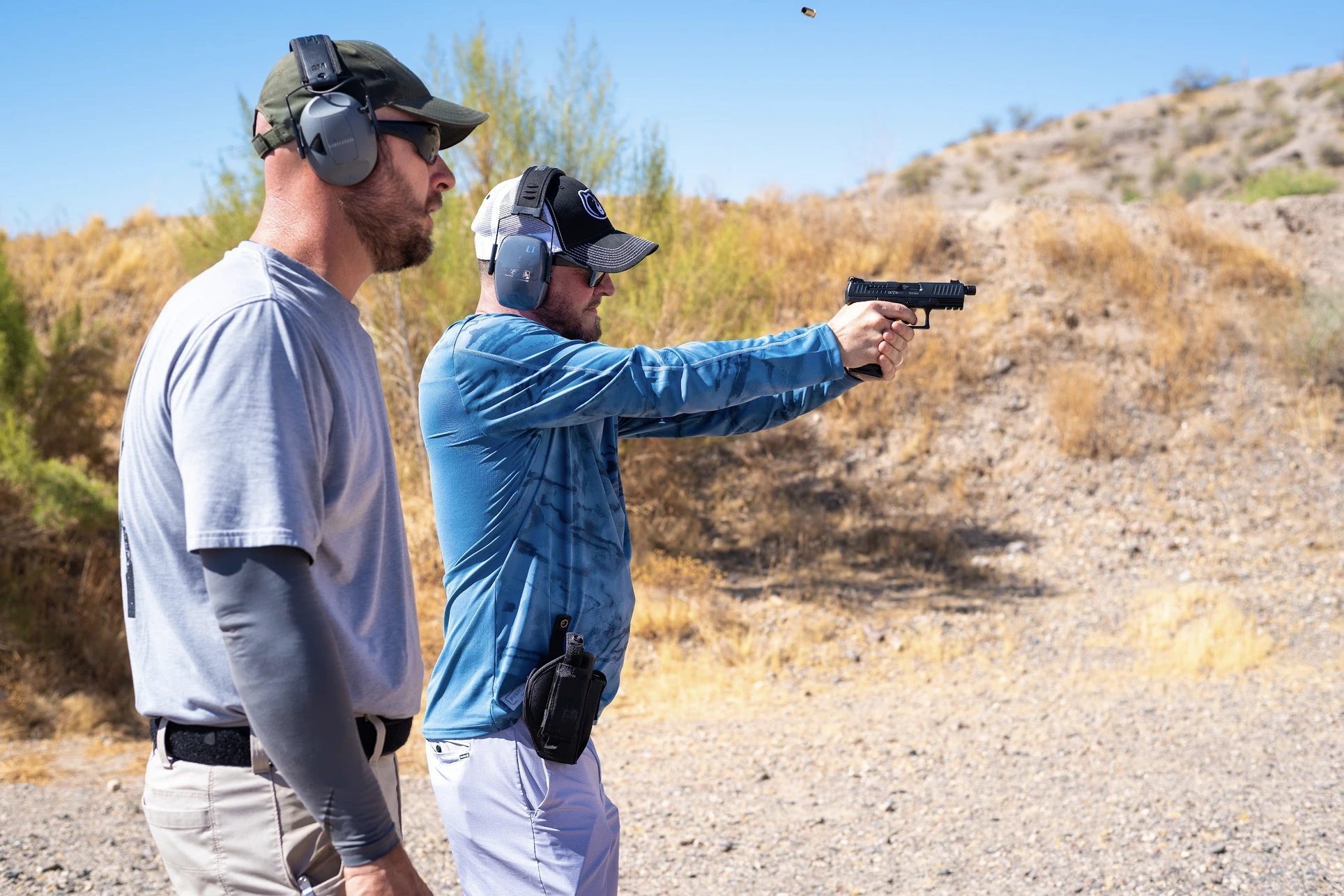 A shooter in a blue long-sleeve performance shirt engages a target downrange with a pistol while an RSO monitors the line; a spent casing is visible mid-air above the slide.