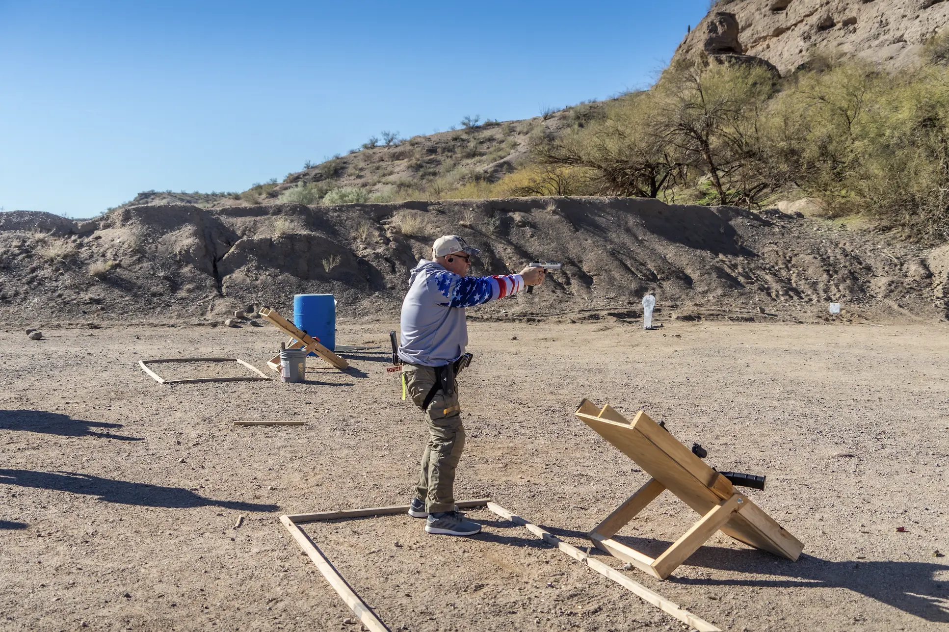 A shooter wearing a patriotic-themed shirt engages a target from within a marked wooden frame in a dusty, high-desert bay. The image captures a real-world training scenario at Cowtown Range.