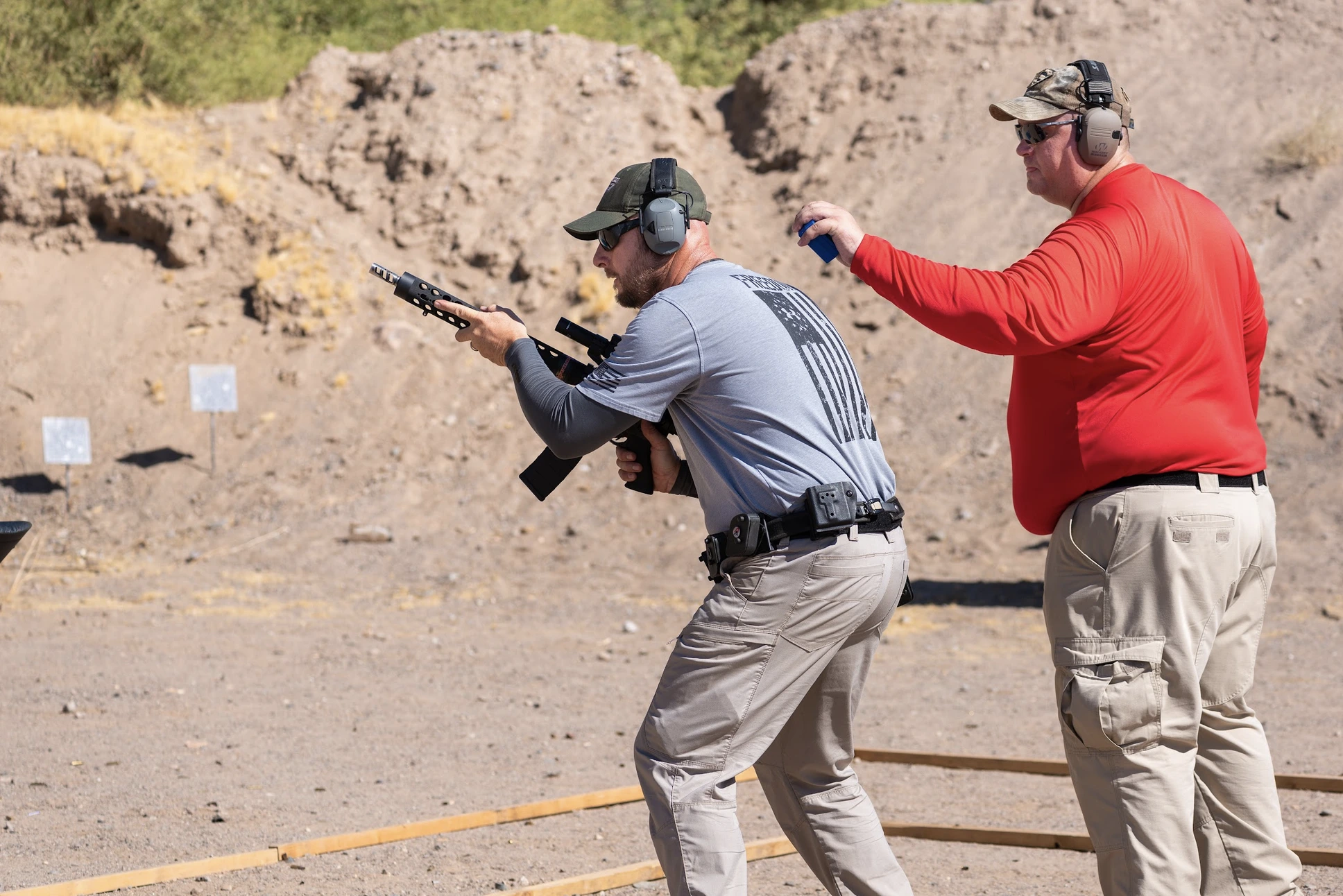 A shooter in tactical gear moves through a course of fire with a rifle platform while a Range Safety Officer in a red shirt follows closely behind with a timer during a training session at Cowtown Range.