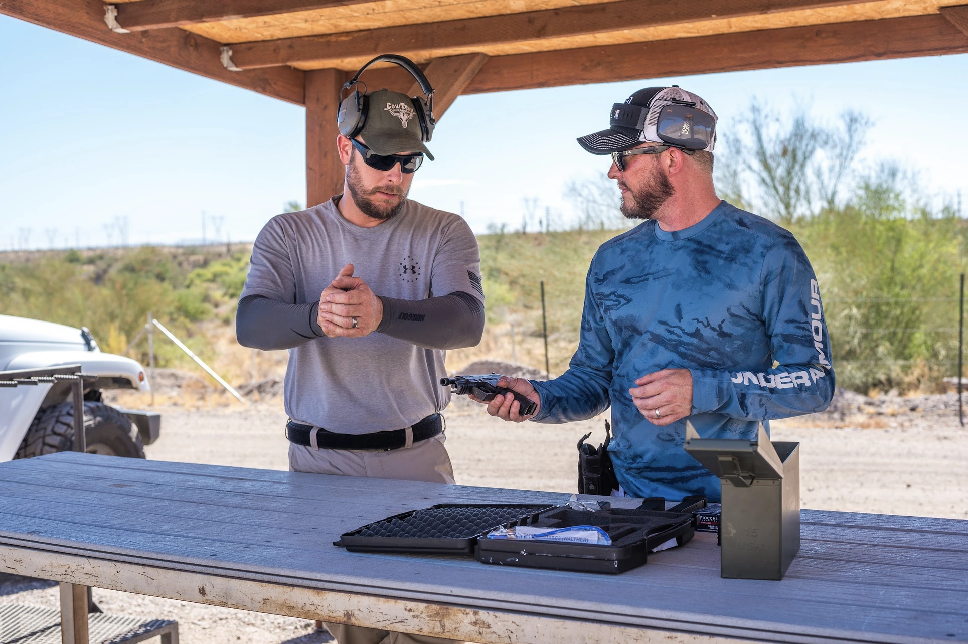 Two shooters engaged in a technical discussion over a handgun system on a wooden range table. This highlights the brand's focus on professional-grade "systems" rather than just recreational shooting.
