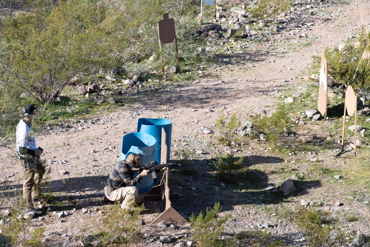A shooter in motion utilizes a blue barrel as a barricade for cover while engaging downrange targets in a dusty, high-desert bay. A mentor stands nearby, providing professional guidance.