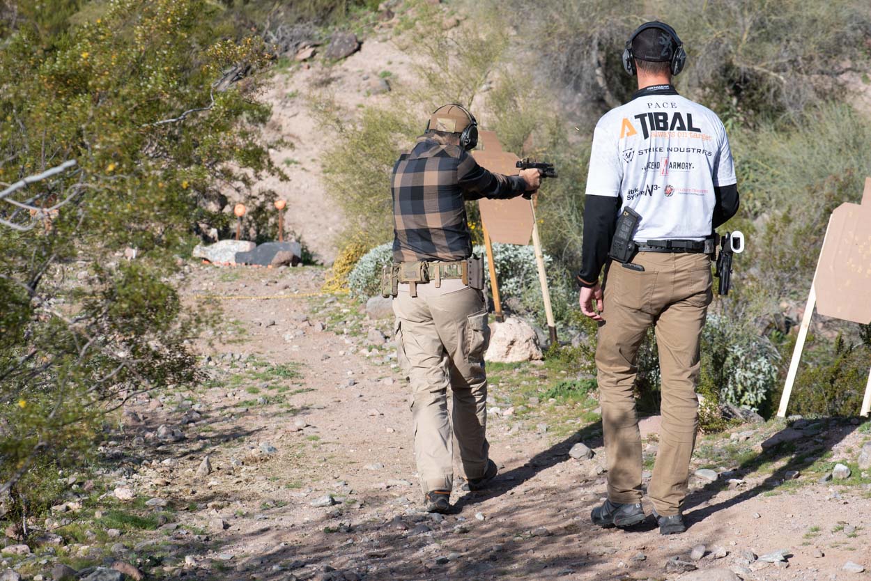 An instructor in a white performance shirt walks alongside a shooter in a flannel shirt and tactical belt. They are moving down a dirt path toward a training scenario with multiple target stands in a rugged, desert environment.