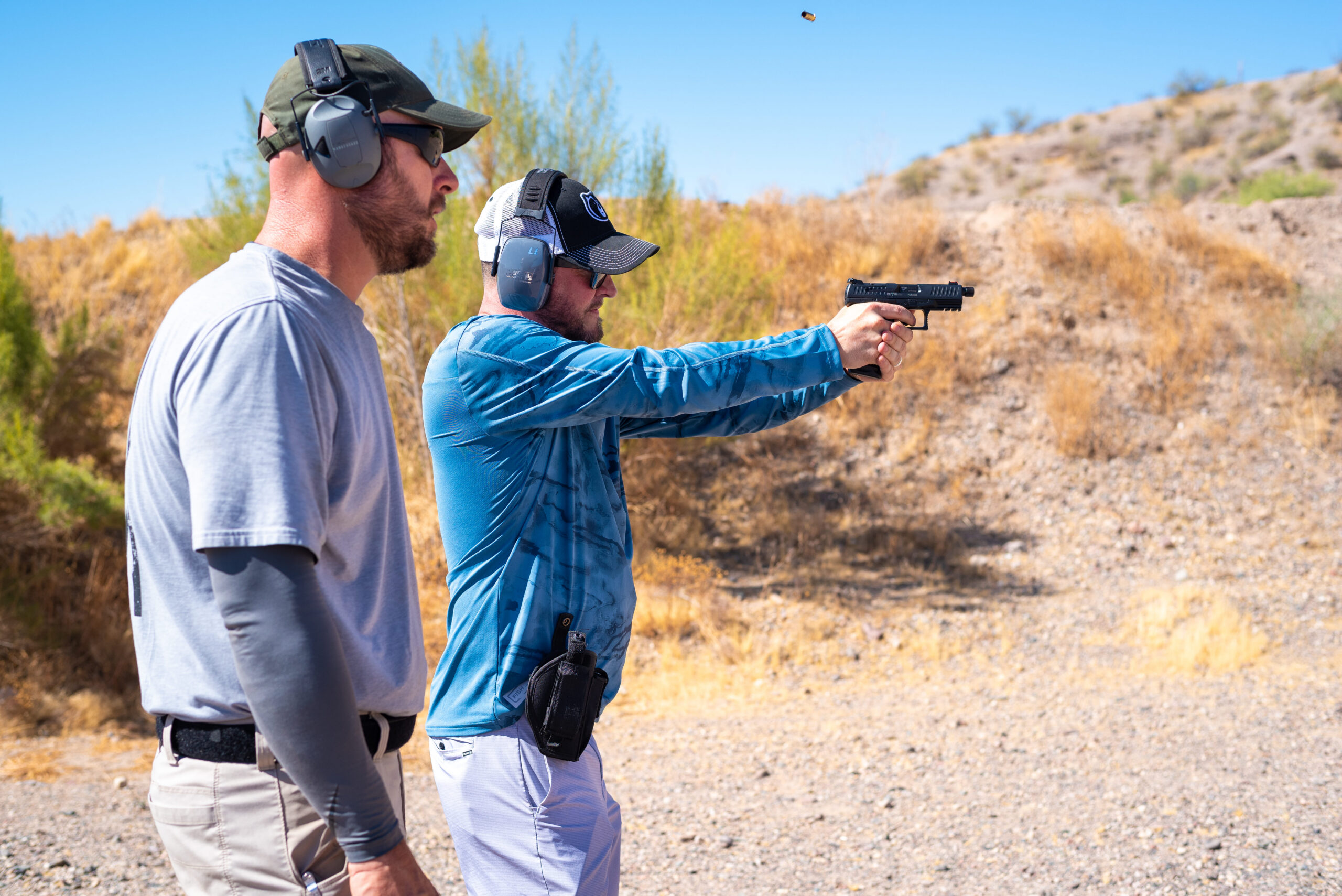 A shooter practicing proper stance and firearm manipulation with a handgun at an outdoor shooting bay under the supervision of an experienced instructor during a tactical training course.