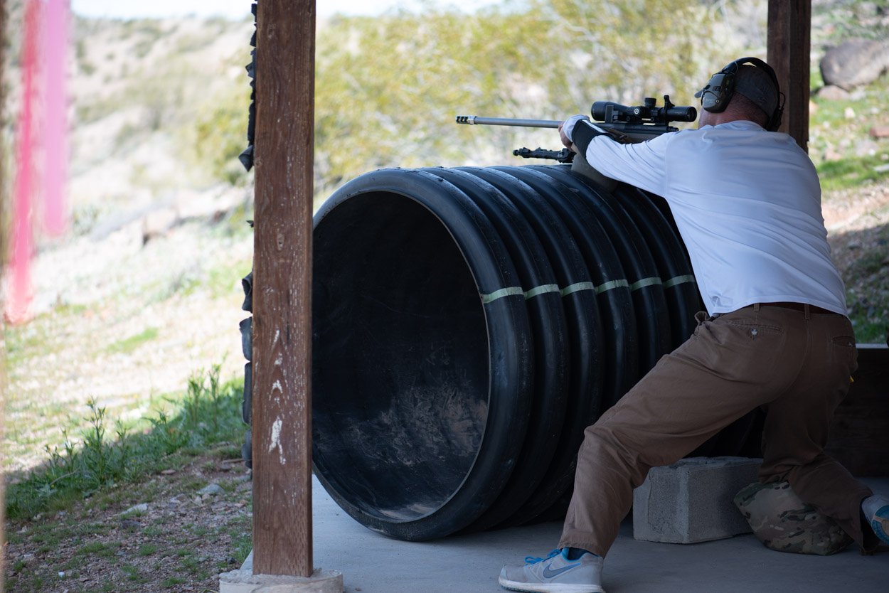 A precision rifle shooter takes a low, stable stance behind a large culvert pipe barricade underneath a shaded outdoor shooting bay in the desert.