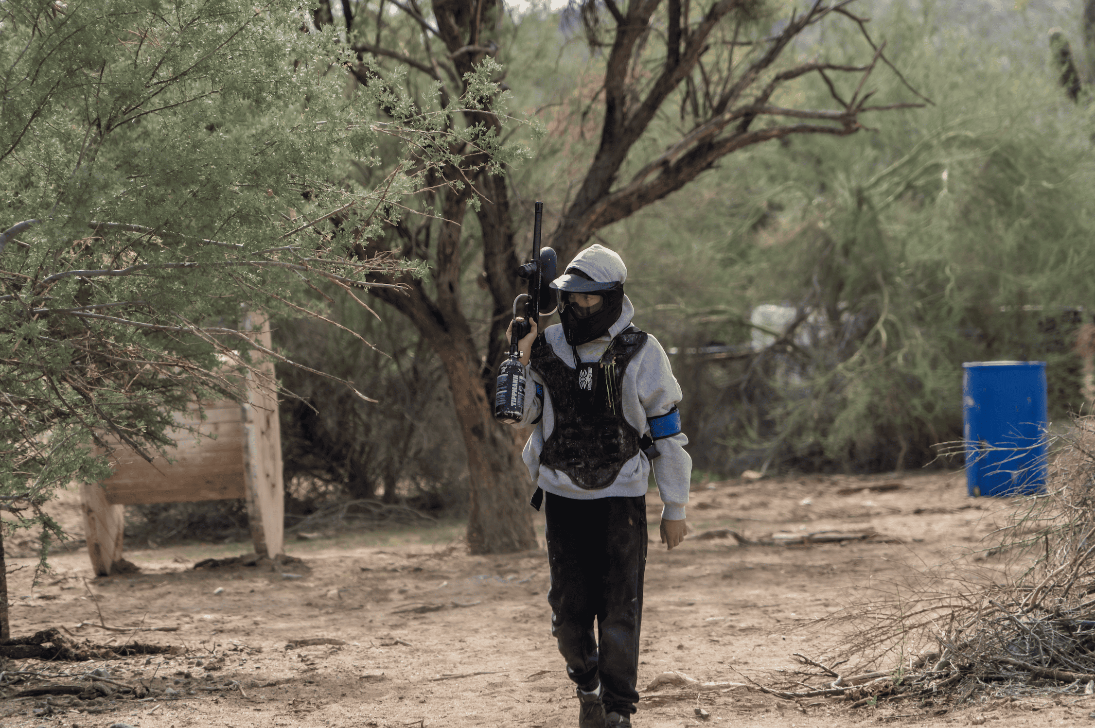 21d37129-ba91-40d1-b205-07199532c114.webp A paintball player wearing a full protective mask, a black tactical chest protector vest, and a gray hoodie walks across the dirt ground at Cowtown Range while carrying a black paintball marker with a Tippmann tank over their shoulder.