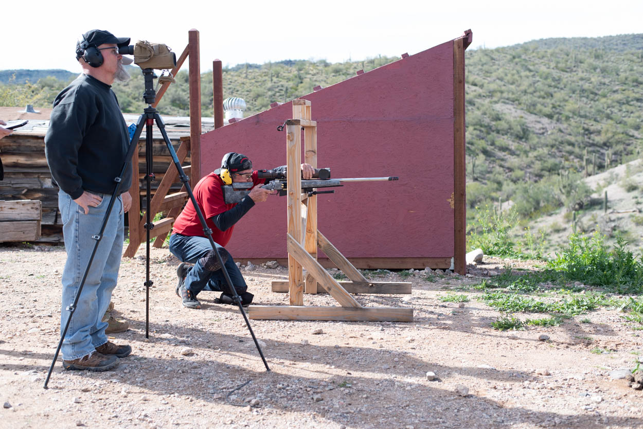 A shooter in a red shirt kneels on a pad, stabilizing a bolt-action precision rifle on a heavy wooden tripod stand to engage a long-range target. Beside him, an observer stands behind a high-magnification spotting scope on a tripod, tracking the impact across a sweeping high-desert valley landscape.