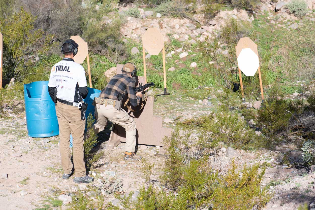 A shooter wearing a flannel shirt and tactical belt stands in a modified crouch, aiming a rifle over a multi-level wooden step barricade. Behind him, an instructor wearing a white jersey branded with "ATIBAL" stands on the dirt path, observing target downrange placement in a desert wash.