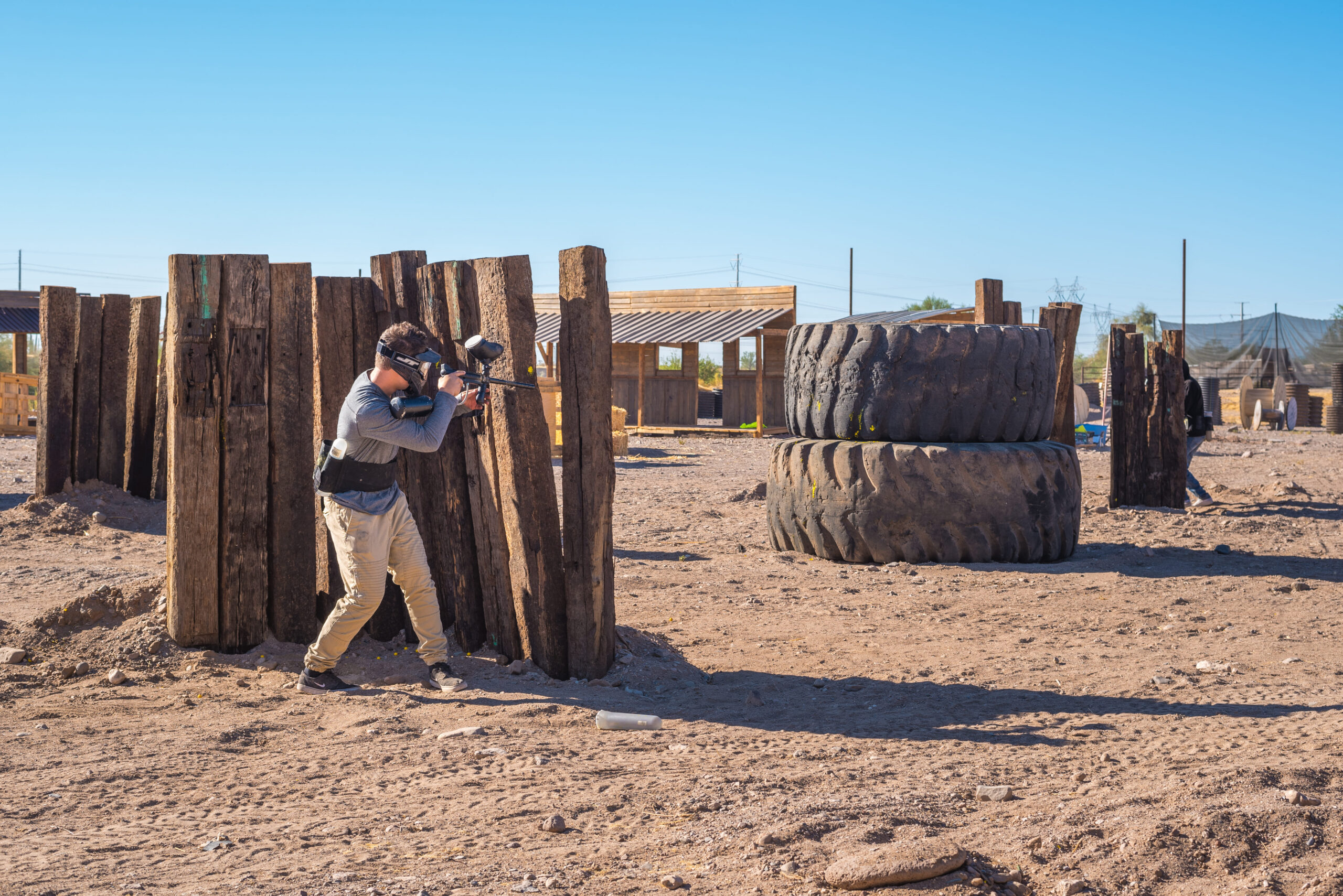 A paintball player in a gray long-sleeve shirt, tan pants, and a protective mask leans out from behind a wooden barricade made of vertical timber posts to aim their marker. The dusty desert outdoor field is scattered with obstacles, including a massive stack of oversized tractor tires in the background.