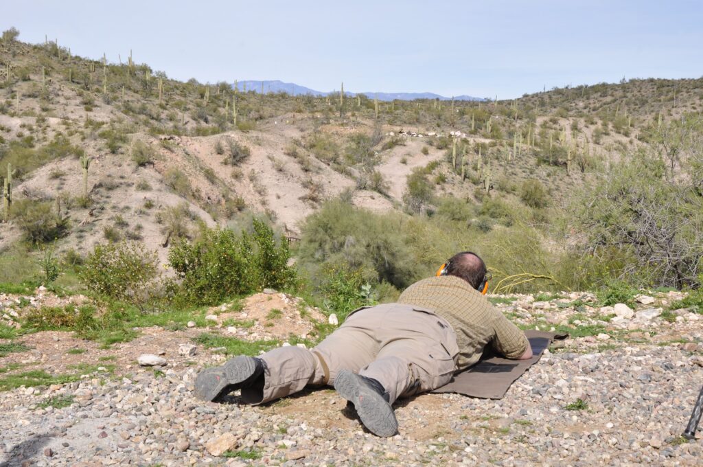 Rear view of a shooter in tactical pants and a plaid shirt practicing long-range shooting from a prone position on a shooting mat, looking out over a wide, rugged Arizona desert canyon filled with saguaros.