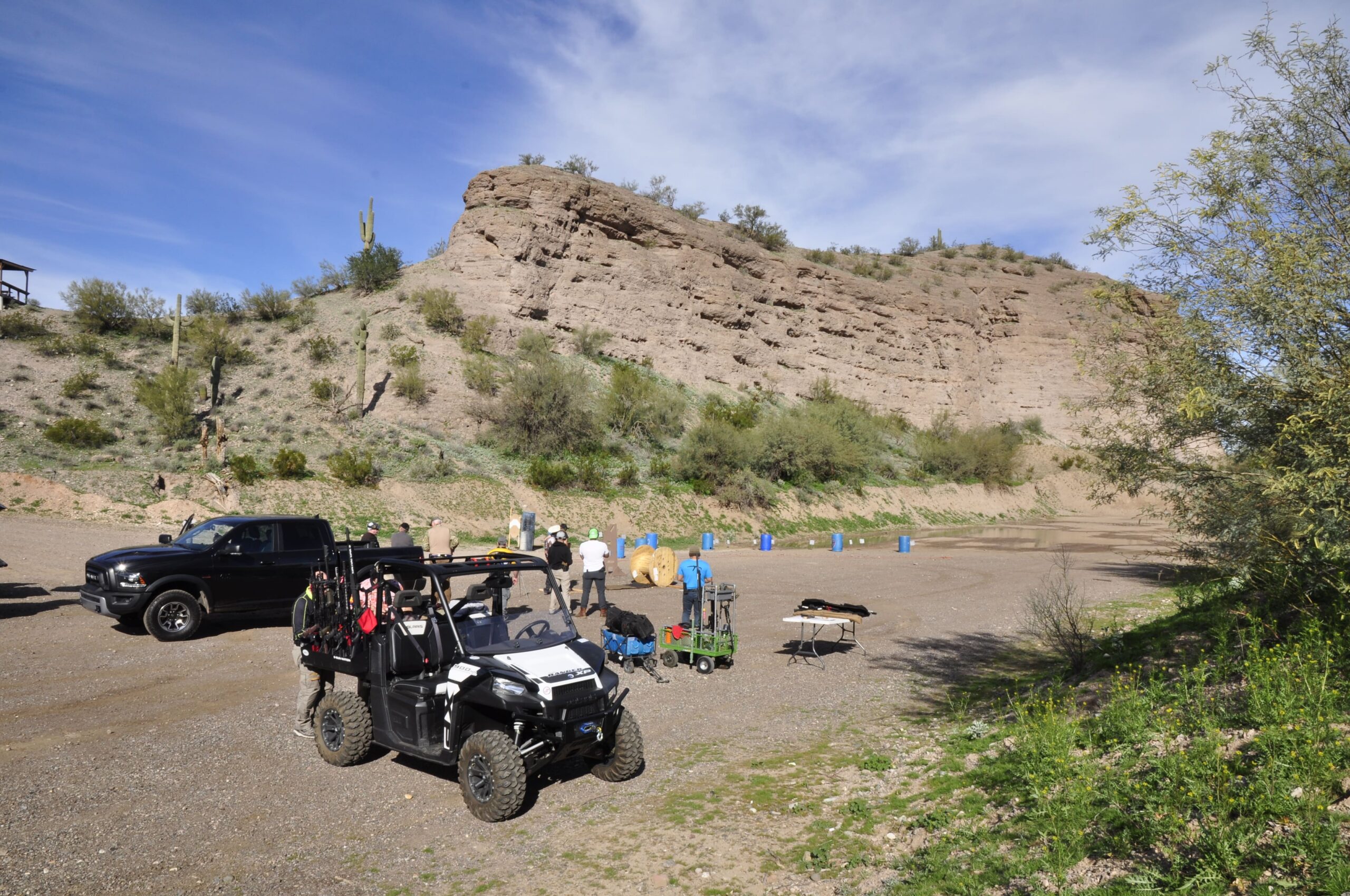 A wide landscape view of an expansive outdoor shooting platform at Cowtown Range. A utility side-by-side vehicle and a black pickup truck are parked on the dirt range flats at the base of a massive, rugged desert hill dotted with saguaro cacti. A small team of operators gathers near target gear and equipment tables under a clear blue sky.