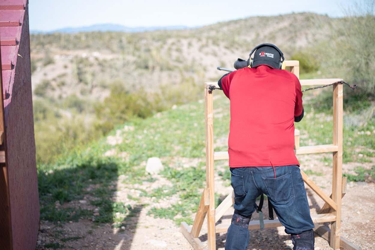 Rear view of a marksman in a red shirt utilizing a wooden stability frame to steady a precision rifle system. The shooter stands overlooking an expansive Arizona desert valley landscape, practicing long-range targeting from an outdoor shooting bay.