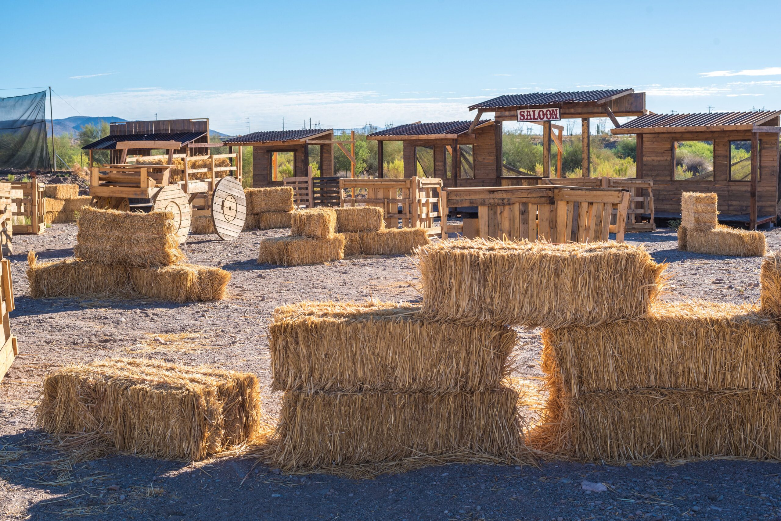A wide landscape shot of an empty 1-acre outdoor paintball field designed like an old Wild West ghost town. The field is densely packed with defensive obstacles made of stacked hay bales, wooden spools, an old cart, and several rustic wooden building facades, including a prominent structure labeled "SALOON."