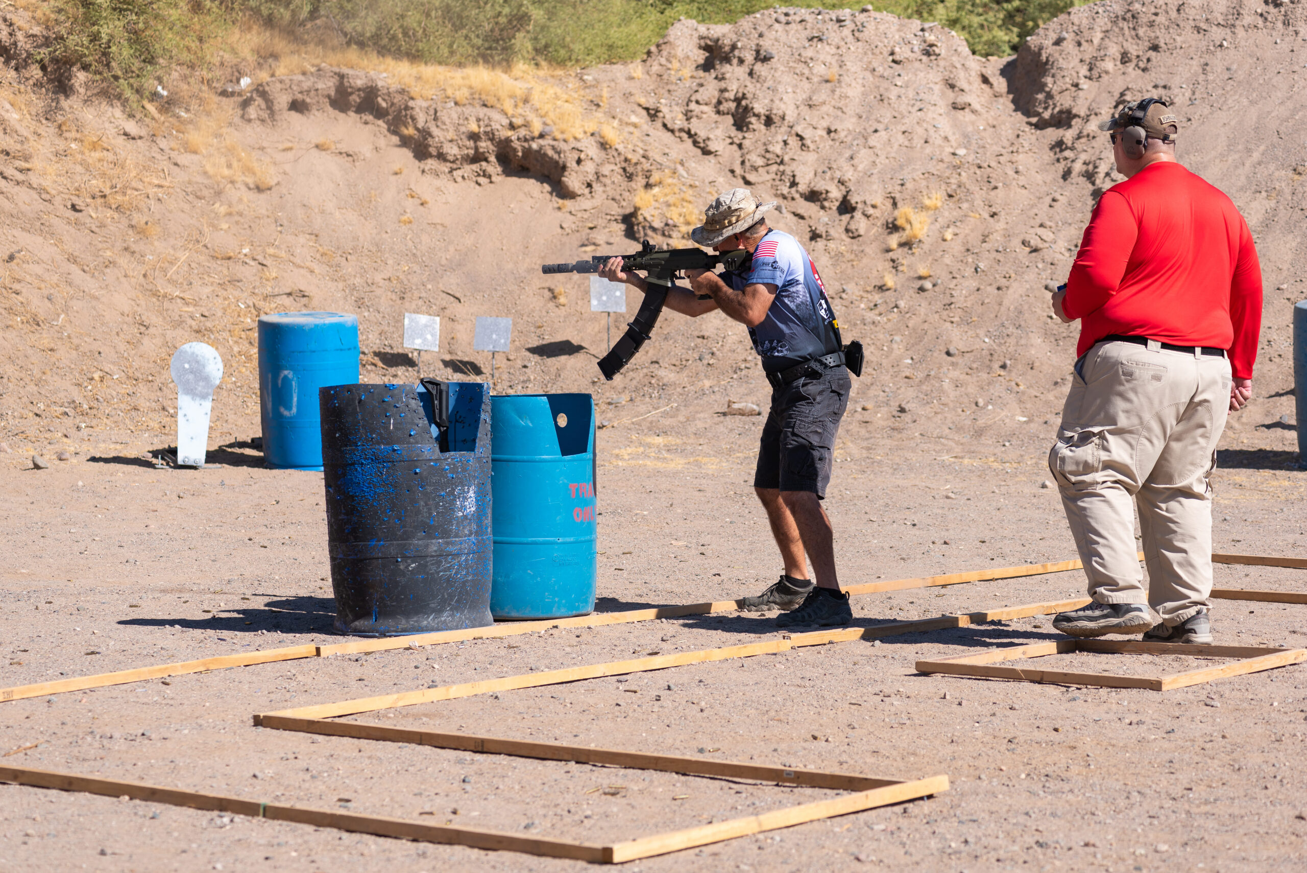 A shooter in a camouflage hat, a short-sleeved tactical jersey, and cargo shorts fires a shotgun while moving through a wood-framed lane boundary. An instructor or safety lead stands nearby on the gravel ground, monitoring the target engagement against a backdrop of a dirt impact berm.