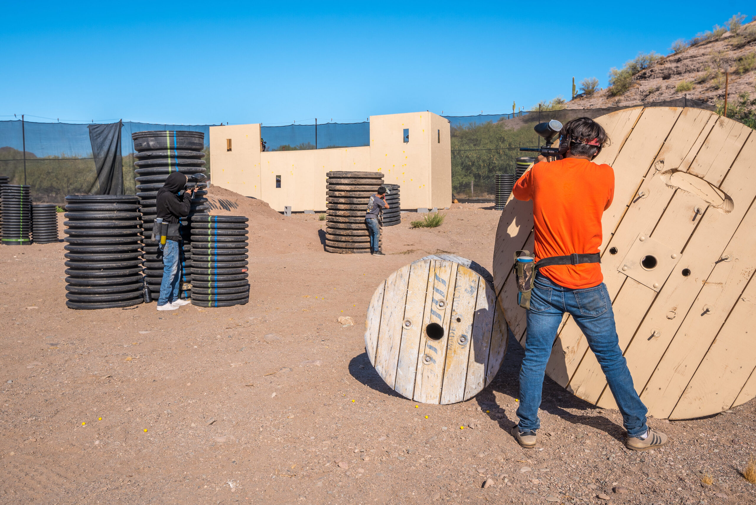 Action shot on an outdoor paintball field featuring players using large wooden cable spools and stacks of black corrugated piping as tactical cover. A player in a bright orange shirt and jeans stands behind a massive leaning wooden spool, holding up a marker on a sunny day with desert foothills in the distance.