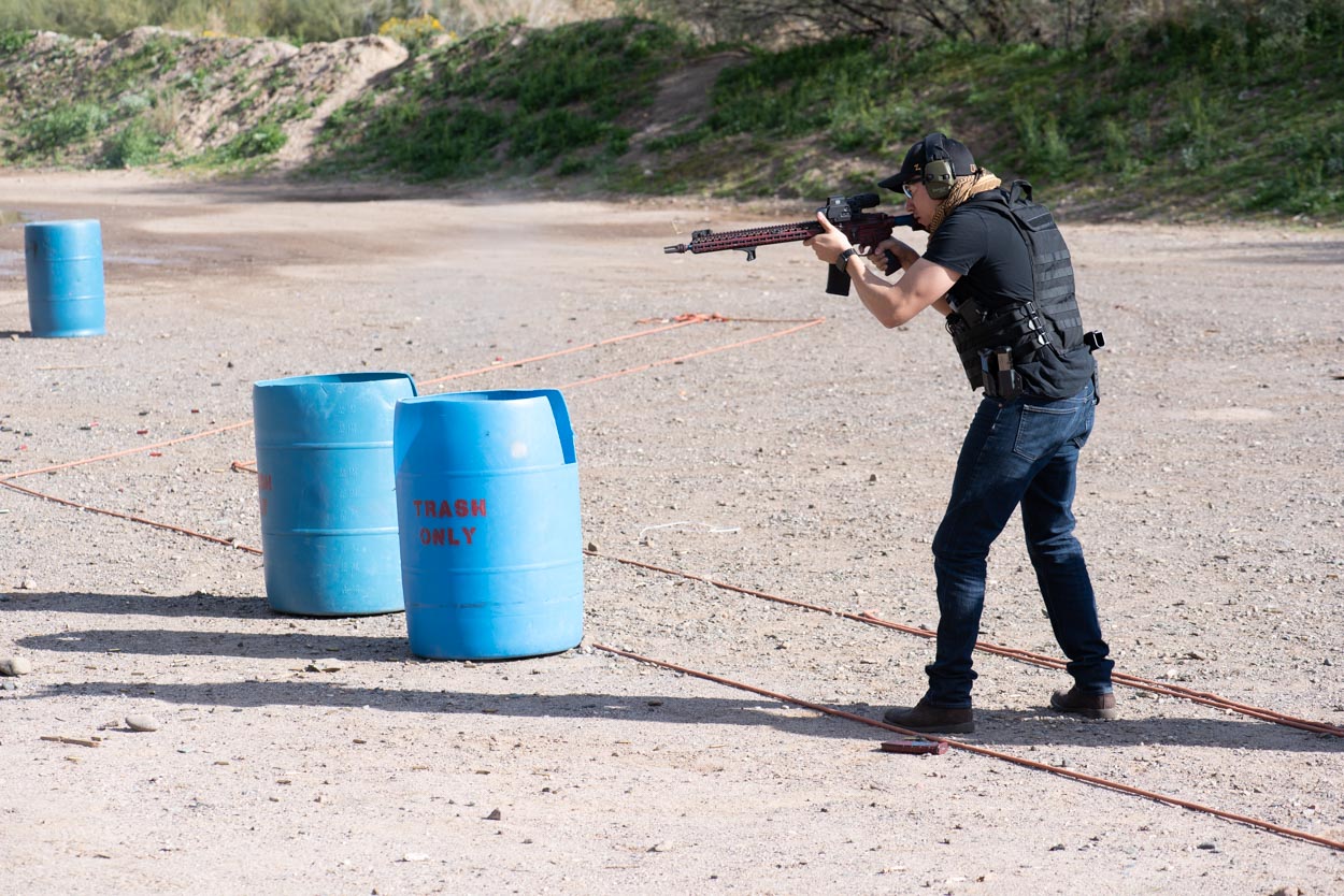 A shooter wearing a black cap, black t-shirt, body armor vest, and jeans stands in a dynamic posture, aiming an AR-15 platform rifle downrange. The desert environment features loose gravel, blue plastic utility barrels labeled trash only, and low hills in the distance.