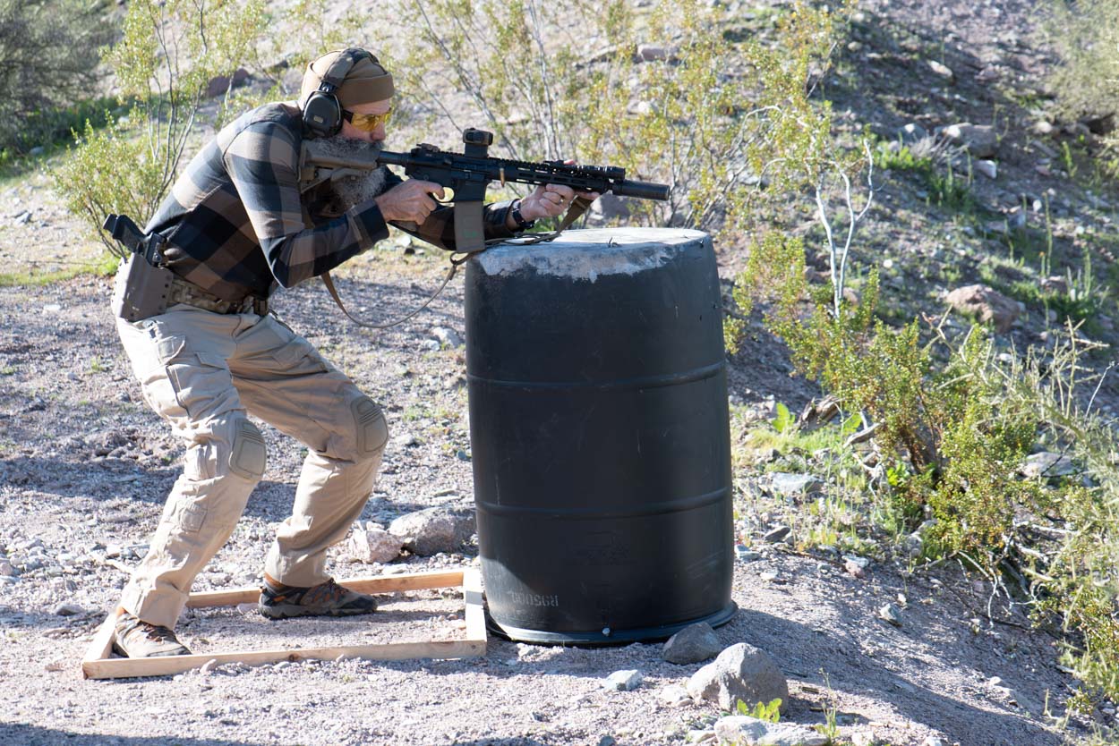 A shooter with a beard and eye protection leans out from behind a large black barrel barricade, actively engaging downrange with a semi-automatic carbine platform equipped with a red dot optic and weapon-mounted light.