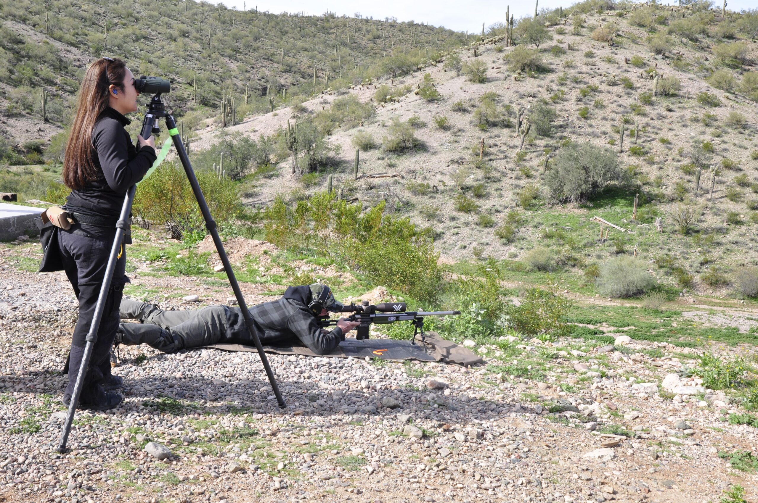 A shooter lies prone on a shooting mat with a precision bolt-action rifle platform on a bipod, while a spotter looks through a tripod-mounted spotting scope looking downrange into a high-desert canyon with saguaro cacti.
