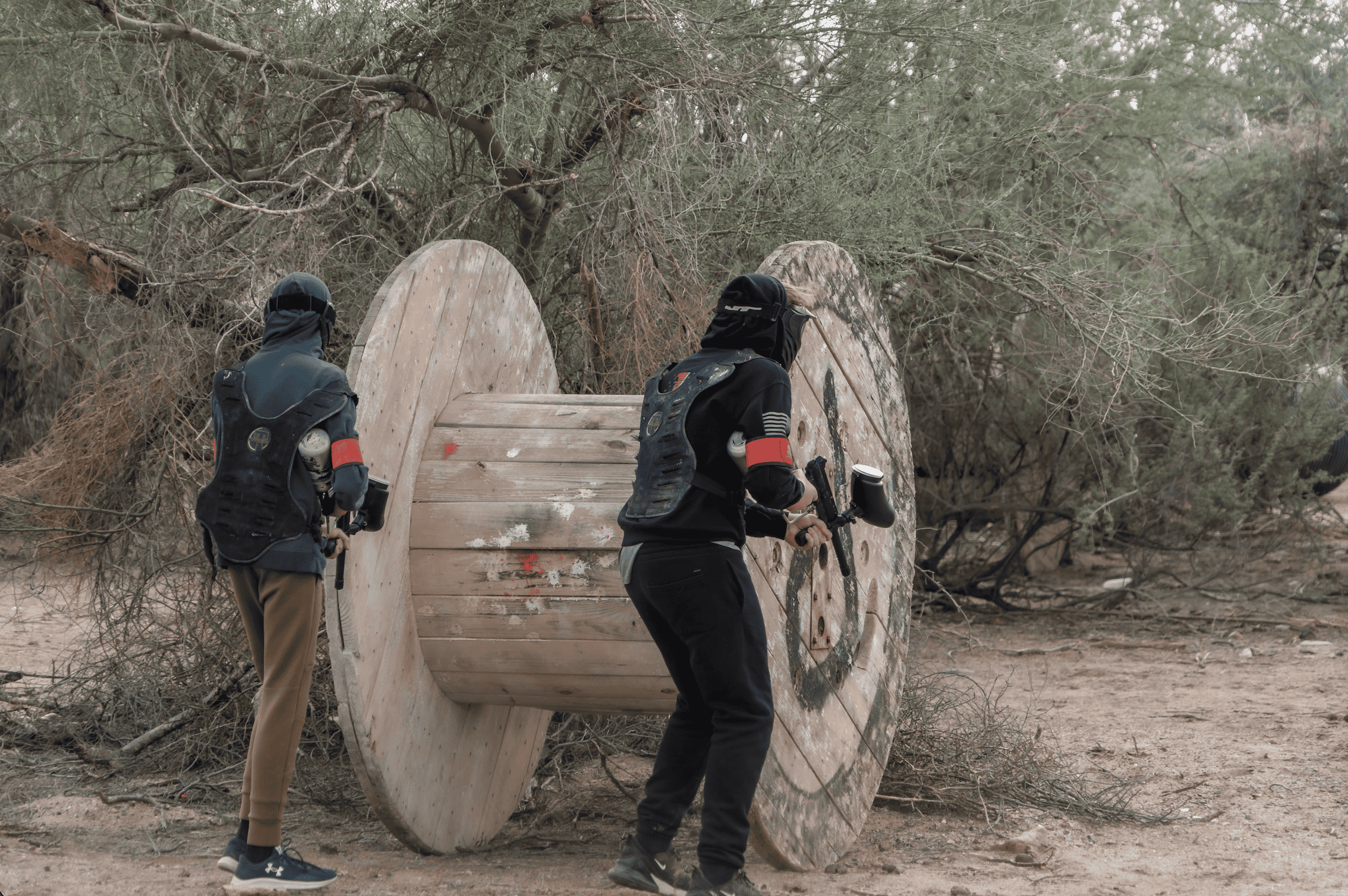 Two paintball players wearing protective masks, body armor vests, and dark hoodies take cover and prepare to advance behind a massive, rustic wooden cable spool on the outdoor desert field at Cowtown Range.