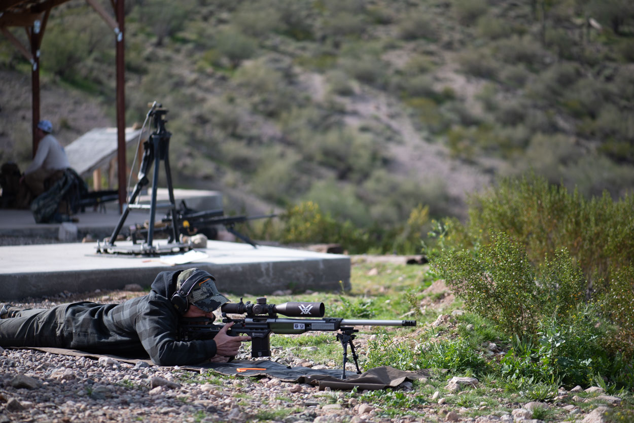 c7f2d02a-c746-4b3a-8658-5f5a2f7f4639.webp A precision rifle shooter lying prone on a shooting mat at an outdoor long-range platform. The shooter is utilizing a bipod-stabilized rifle system equipped with a high-magnification optic to scan and engage distant targets across a dusty, desert hillside backdrop.