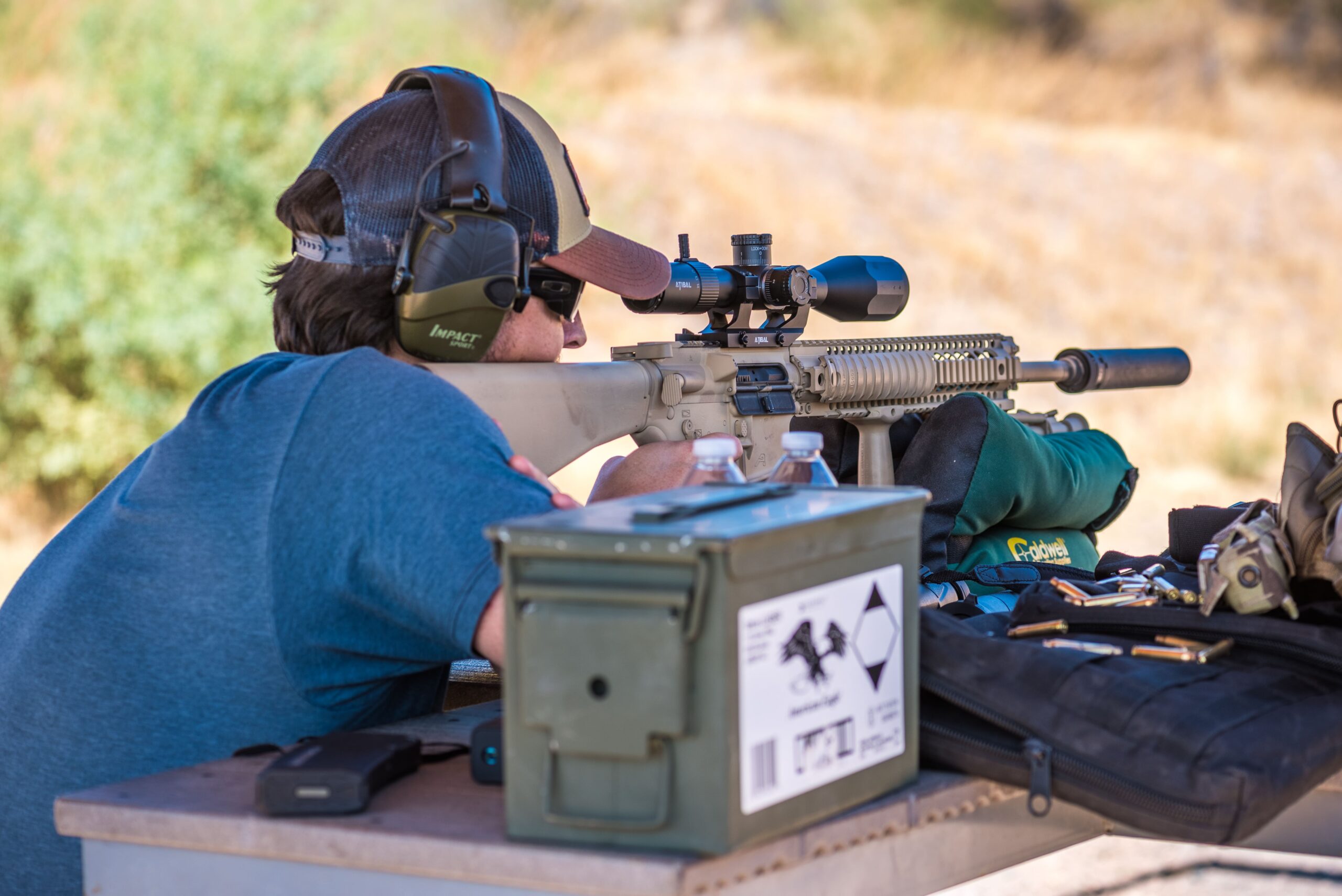 da865b6e-29d1-4752-a659-f3e9ac94a59c.webp A marksman wearing ear protection utilizes a shooting bag on a bench to stabilize an accurized AR rifle platform equipped with a high-magnification optics system at an outdoor range bay.