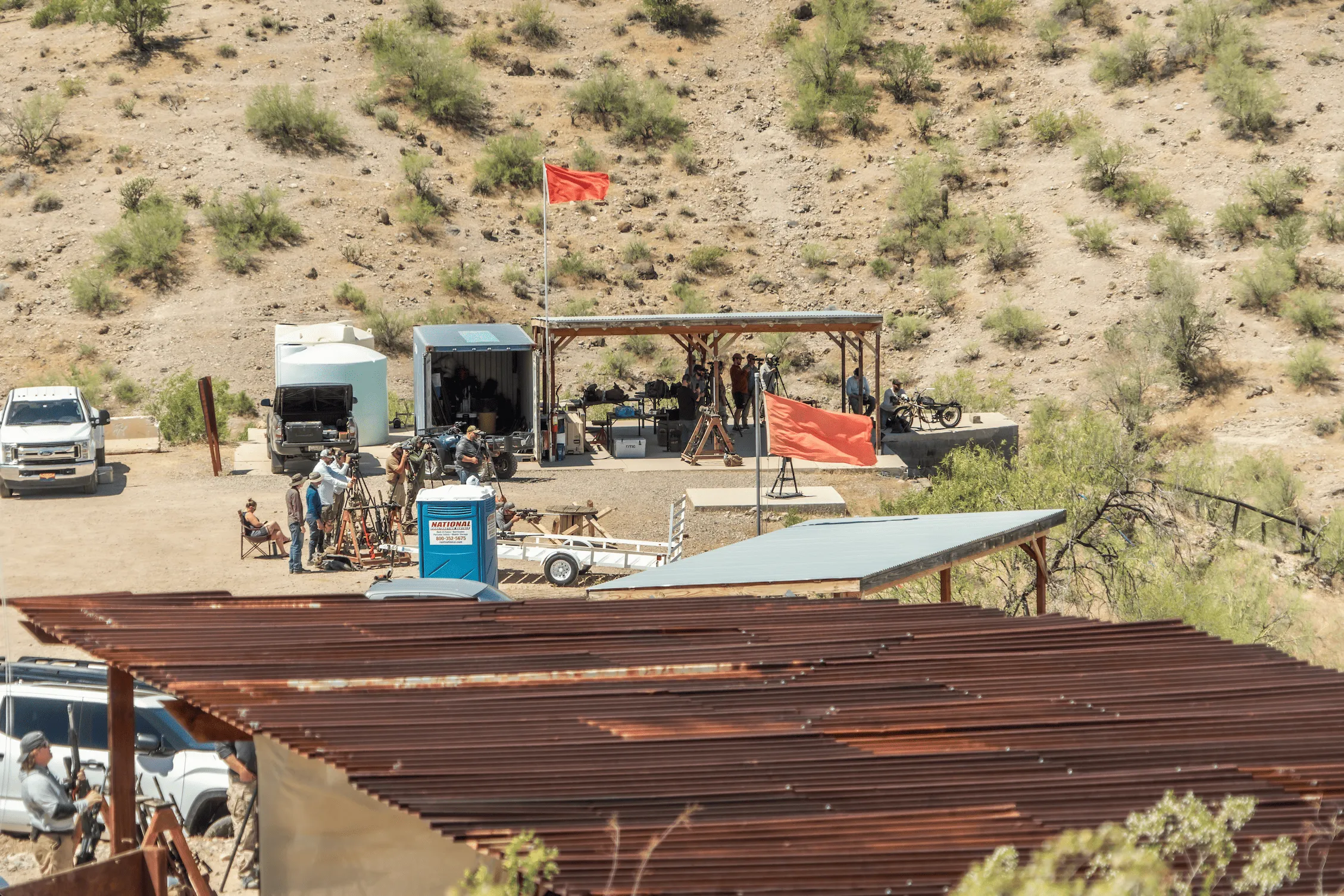 High-angle view of an outdoor desert shooting range with shade structures, vehicles, and active shooters.