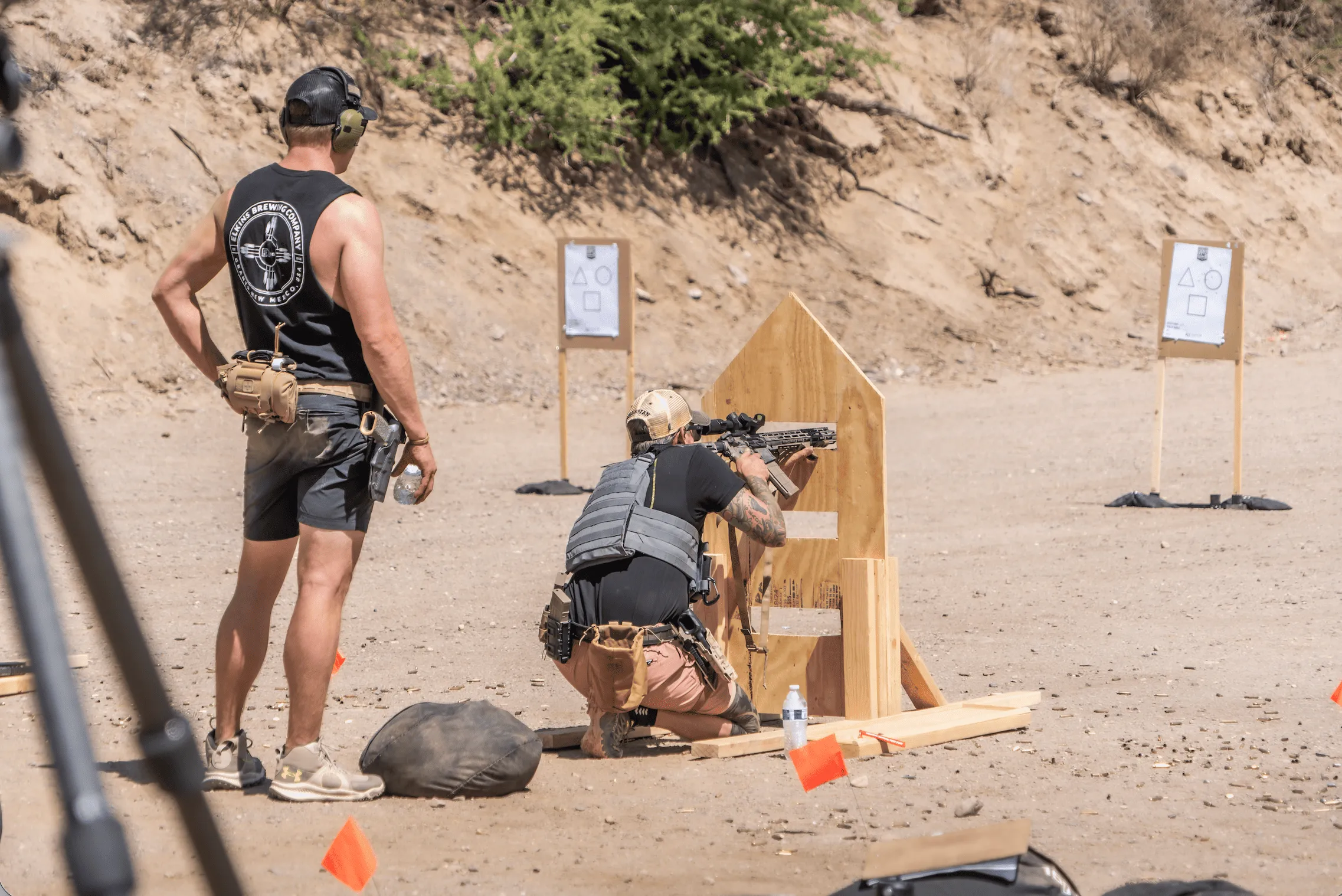 An instructor monitors a shooter practicing best practices for carbine handling while executing a kneeling barricade drill at an outdoor shooting bay.