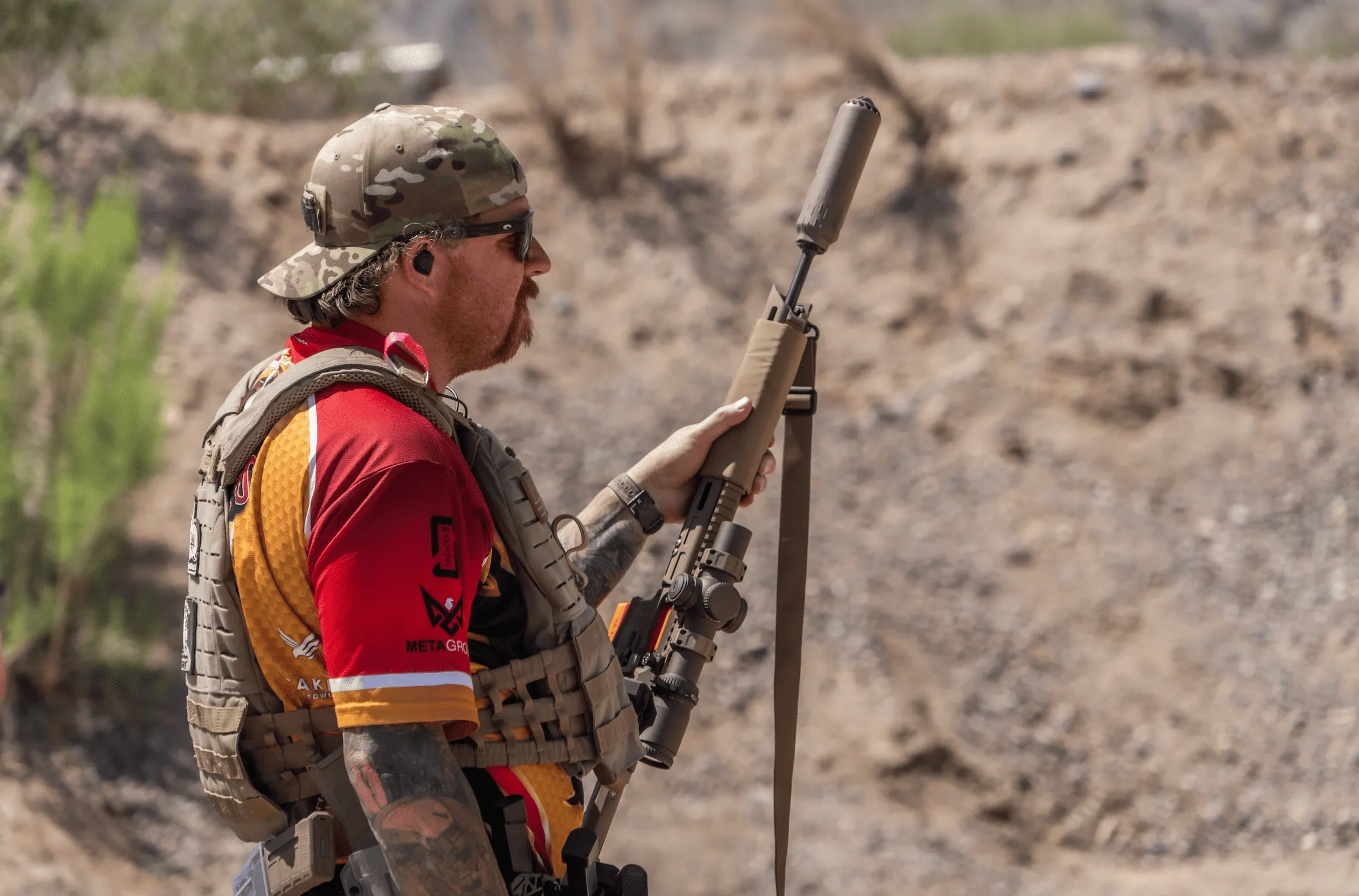 A tactical shooter in a camo hat, sunglasses, and chest rig holds a suppressed carbine platform equipped with a low-power variable optic (LPVO) while practicing safe muzzle discipline at a high-desert outdoor range bay.