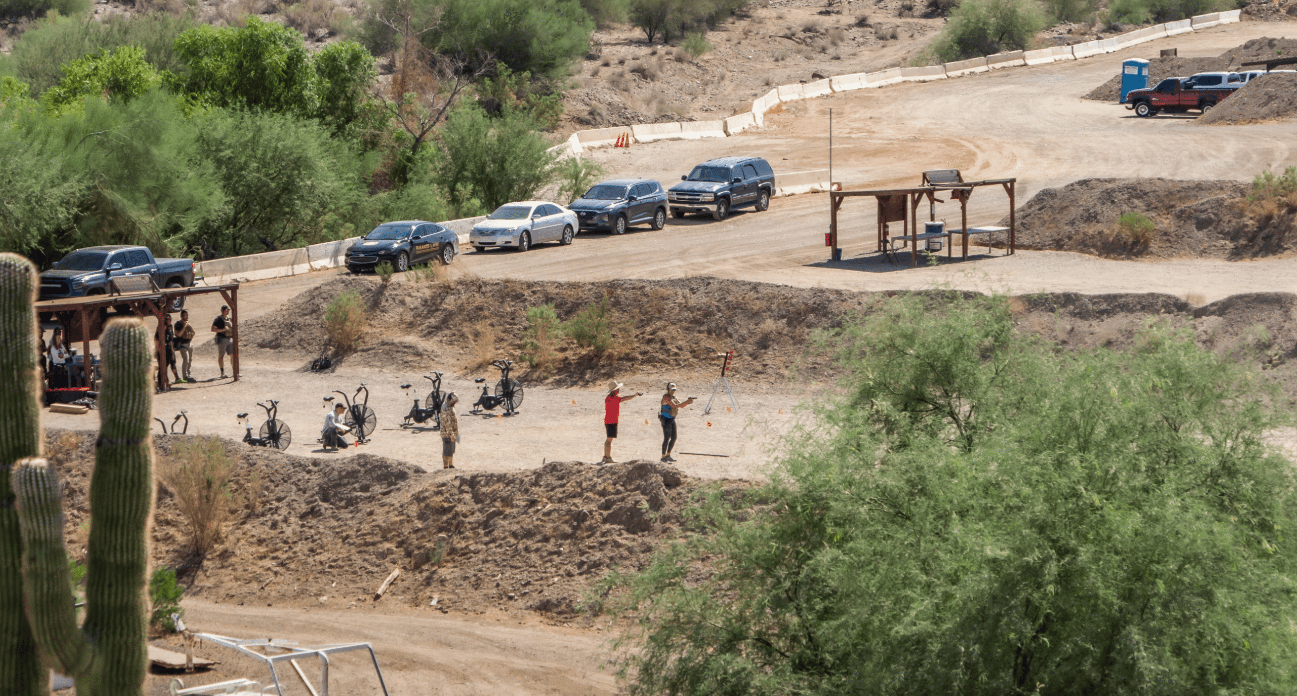 1048ccef-69b8-447f-9736-436914f8bee4.webp Elevated wide shot of an outdoor tactical shooting bay at Cowtown Range in Arizona, showing multiple shooters training with stationary exercise bikes and targets, with parked vehicles lined up on a dirt path in the high-desert landscape.