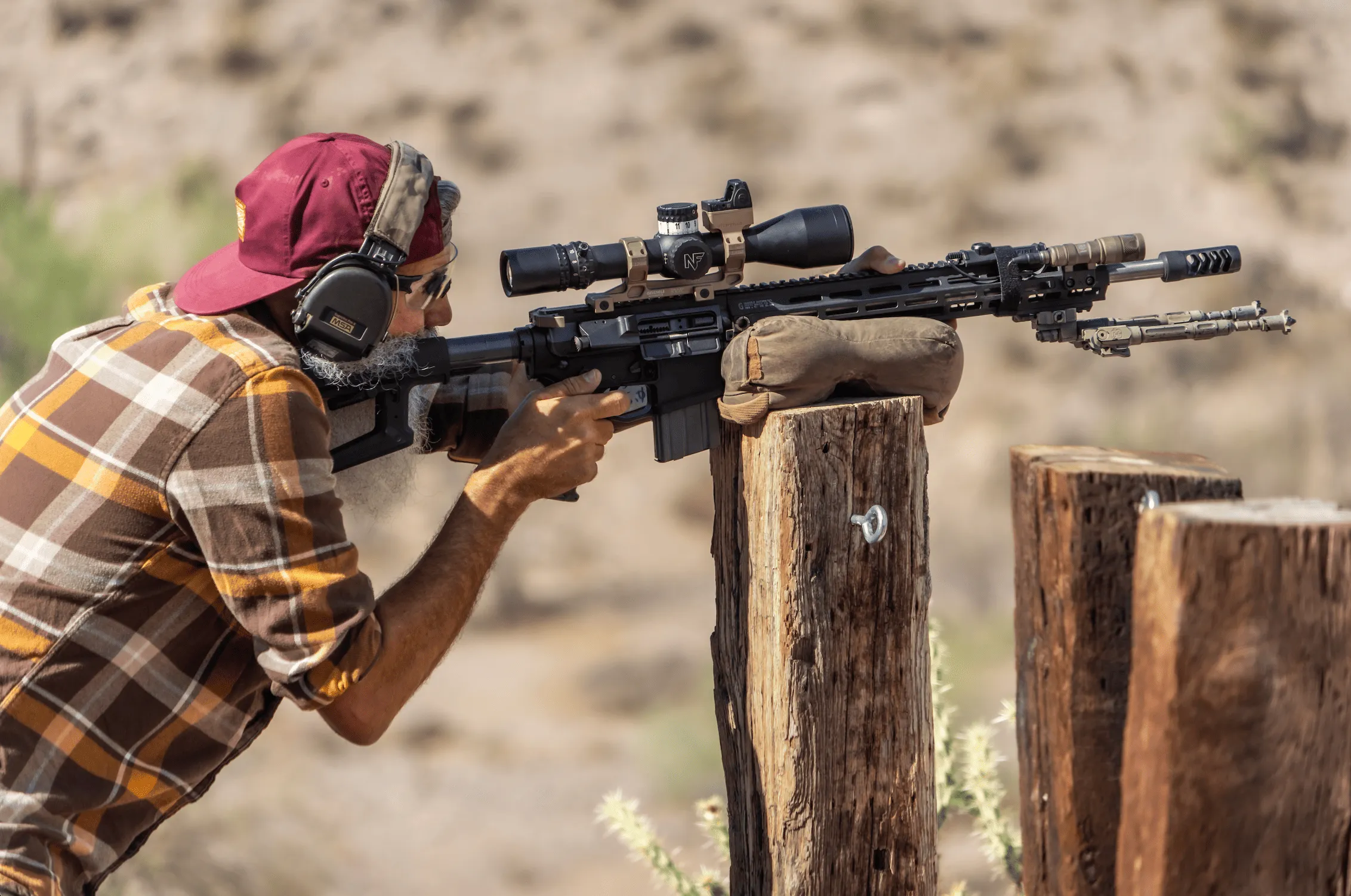 A shooter with a grey beard aims a scoped rifle rested on a sandbag over a wooden post in the desert.