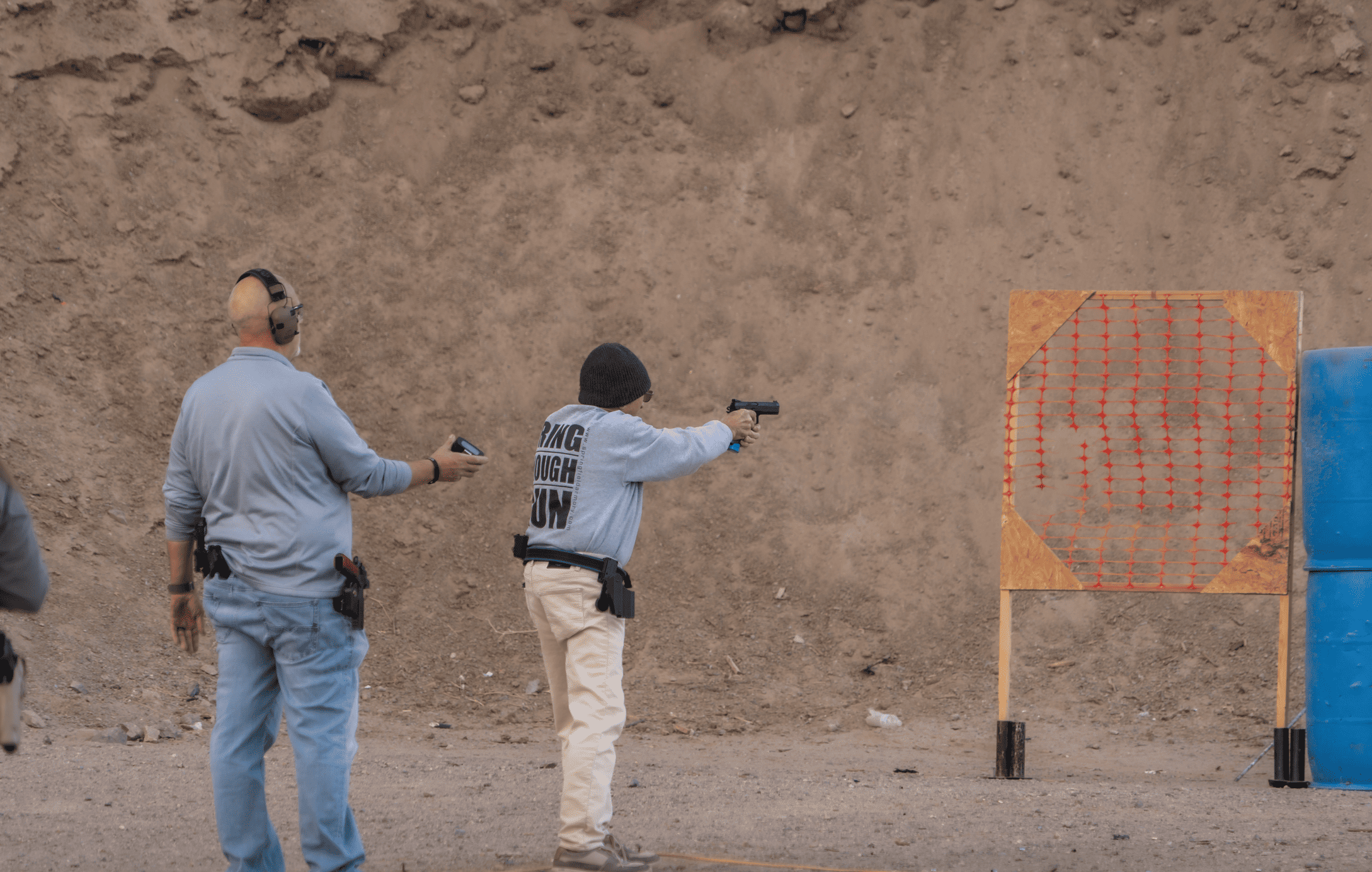 An outdoor training scenario showing a range officer holding a shot timer next to a shooter in a black beanie and a grey sweatshirt who is aiming a handgun at an orange mesh target stand.