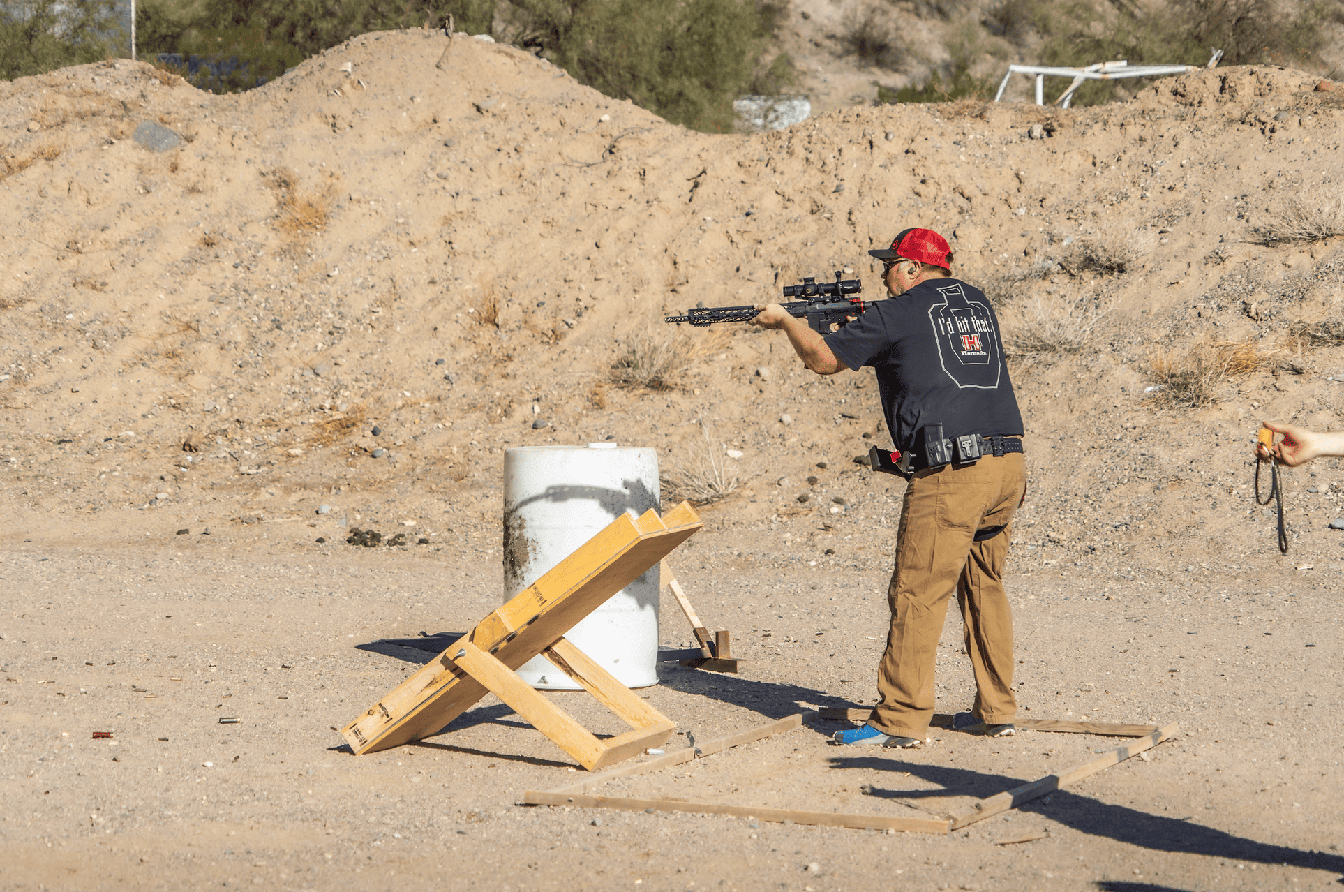 A male shooter in a black t-shirt and tan tactical pants stands in a designated outdoor shooting bay at Cowtown Range, actively aiming a black rifle system equipped with an optic during a dynamic training drill.