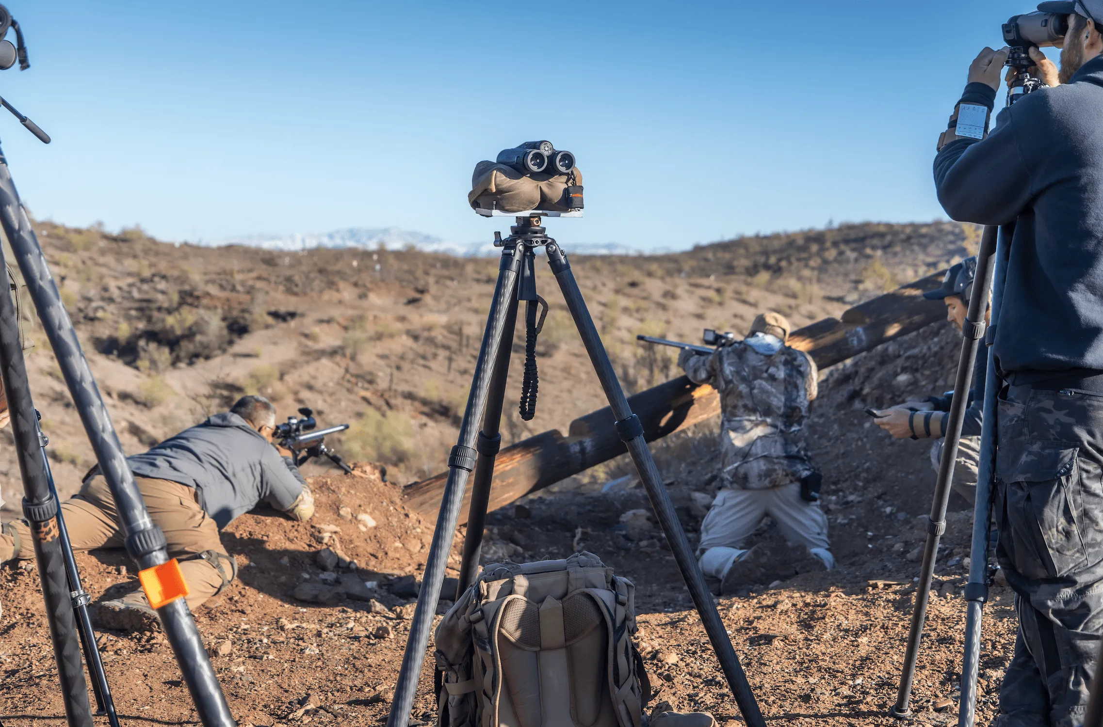 Experienced shooters and spotters running dynamic long-range drills on a rugged desert hillside. One shooter lies prone behind a precision rifle while another kneels using a large log barrier, supported by spotters using tripod-mounted optics to track impacts downrange.
