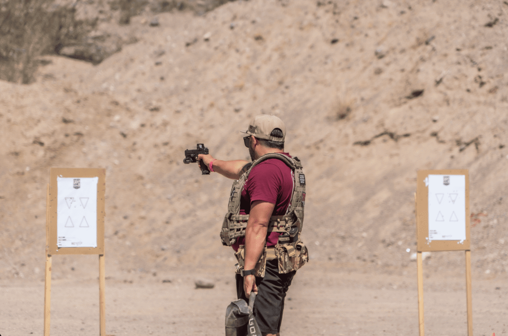 An outdoor, medium-shot of a shooter wearing a maroon shirt, a tan baseball cap, and a tactical plate carrier vest, aiming a handgun equipped with an optic at a paper training target inside a high-desert bay.