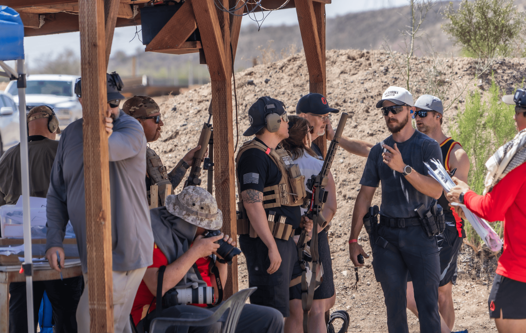 Shooters and range officers wearing plate carriers, baseball caps, and sunglasses gather under a rugged wooden awning on a desert range to review paperwork and firearms. A photographer with a massive camera lens sits in the foreground capturing the action.
