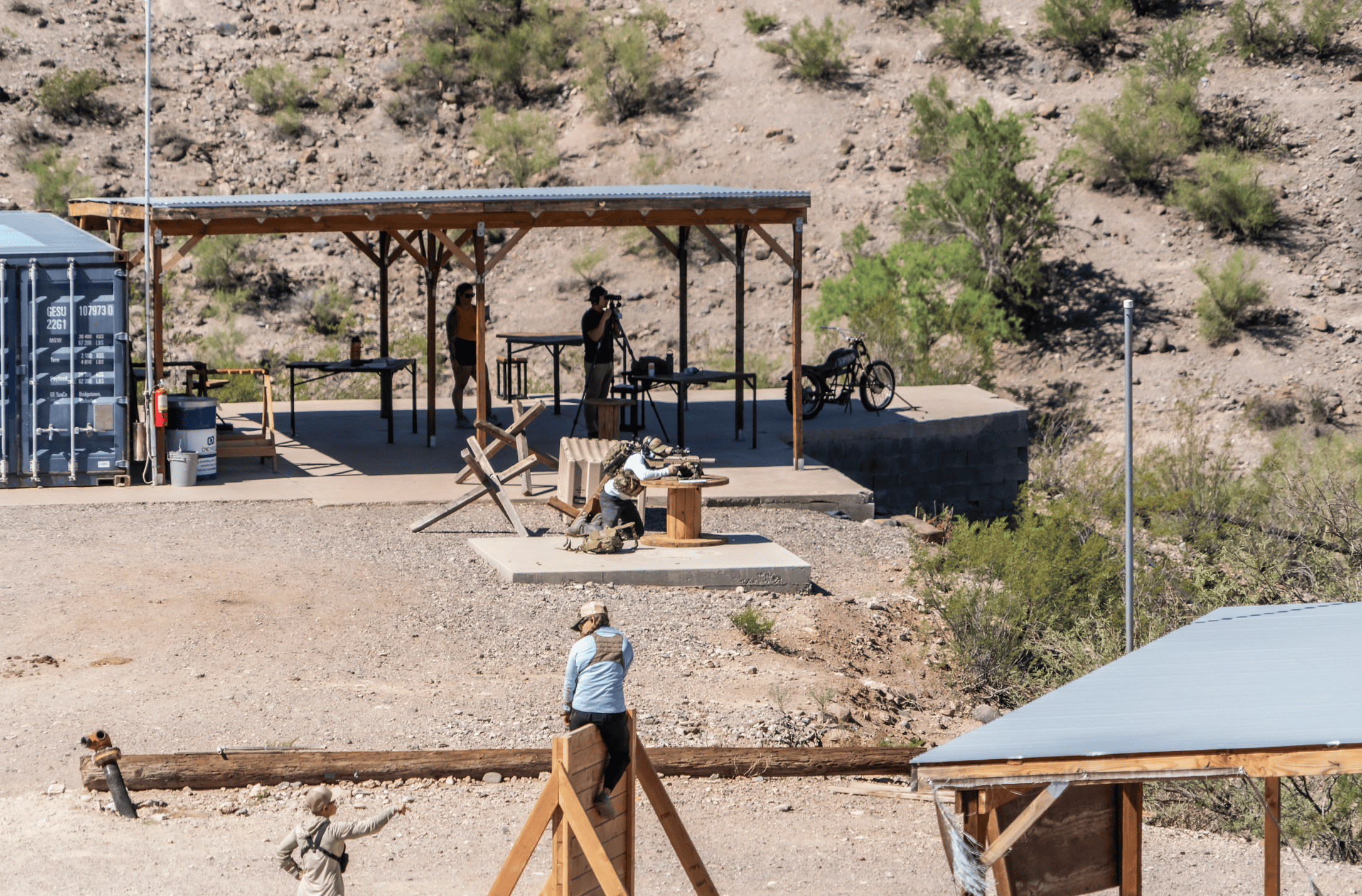 Shooters engaging in long-range precision and tactical drills under a shaded wooden awning platform at an outdoor desert shooting facility.