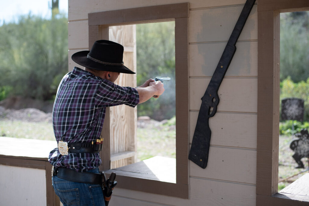 A Cowboy Action Shooter in a leather hat, bandana, and historical western shirt firing a single-action revolver one-handed out of a wooden shack window, surrounded by a thick cloud of white gunsmoke.