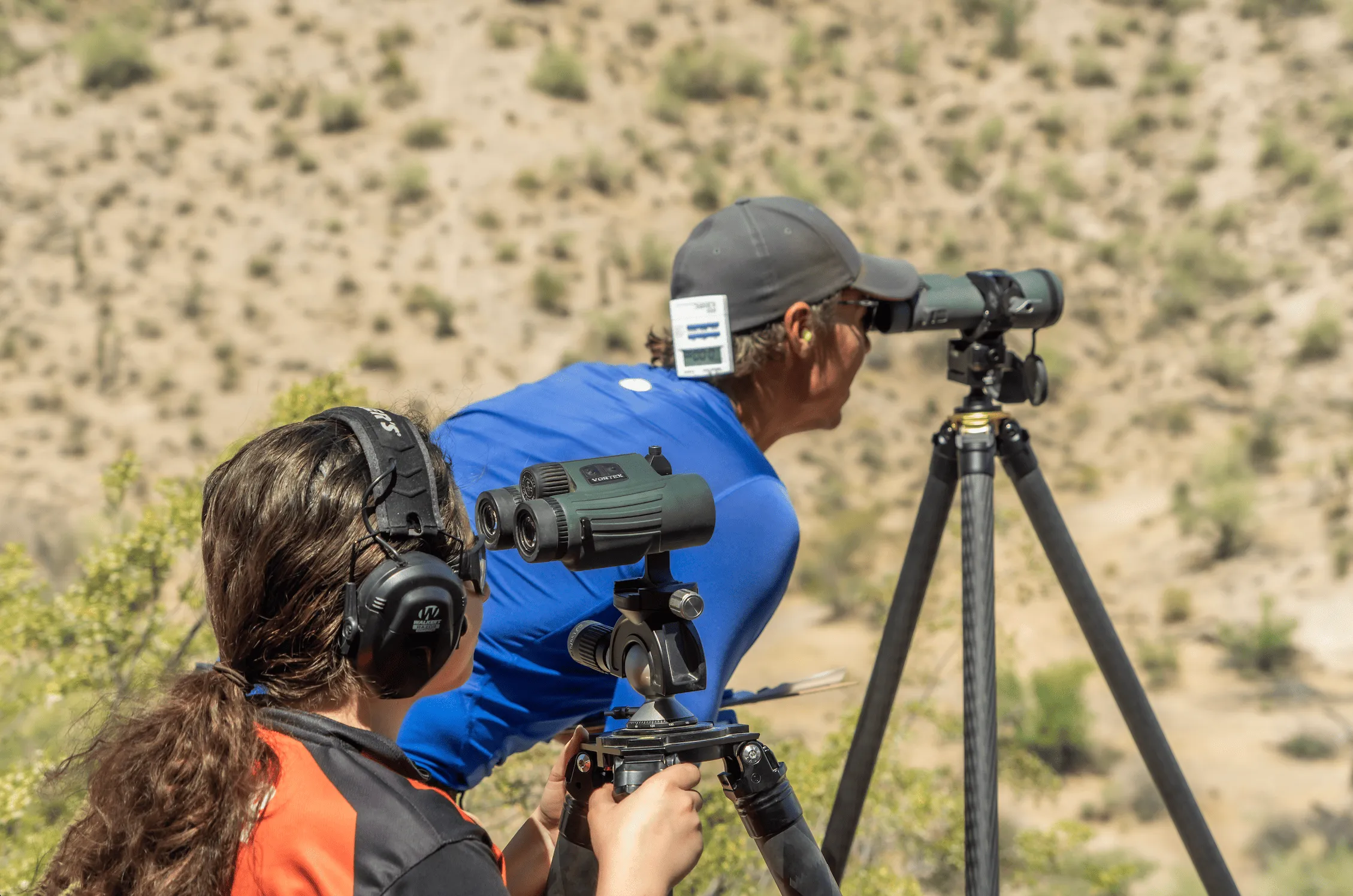 Two shooters using a spotting scope and binoculars on tripods to observe downrange in a desert landscape.