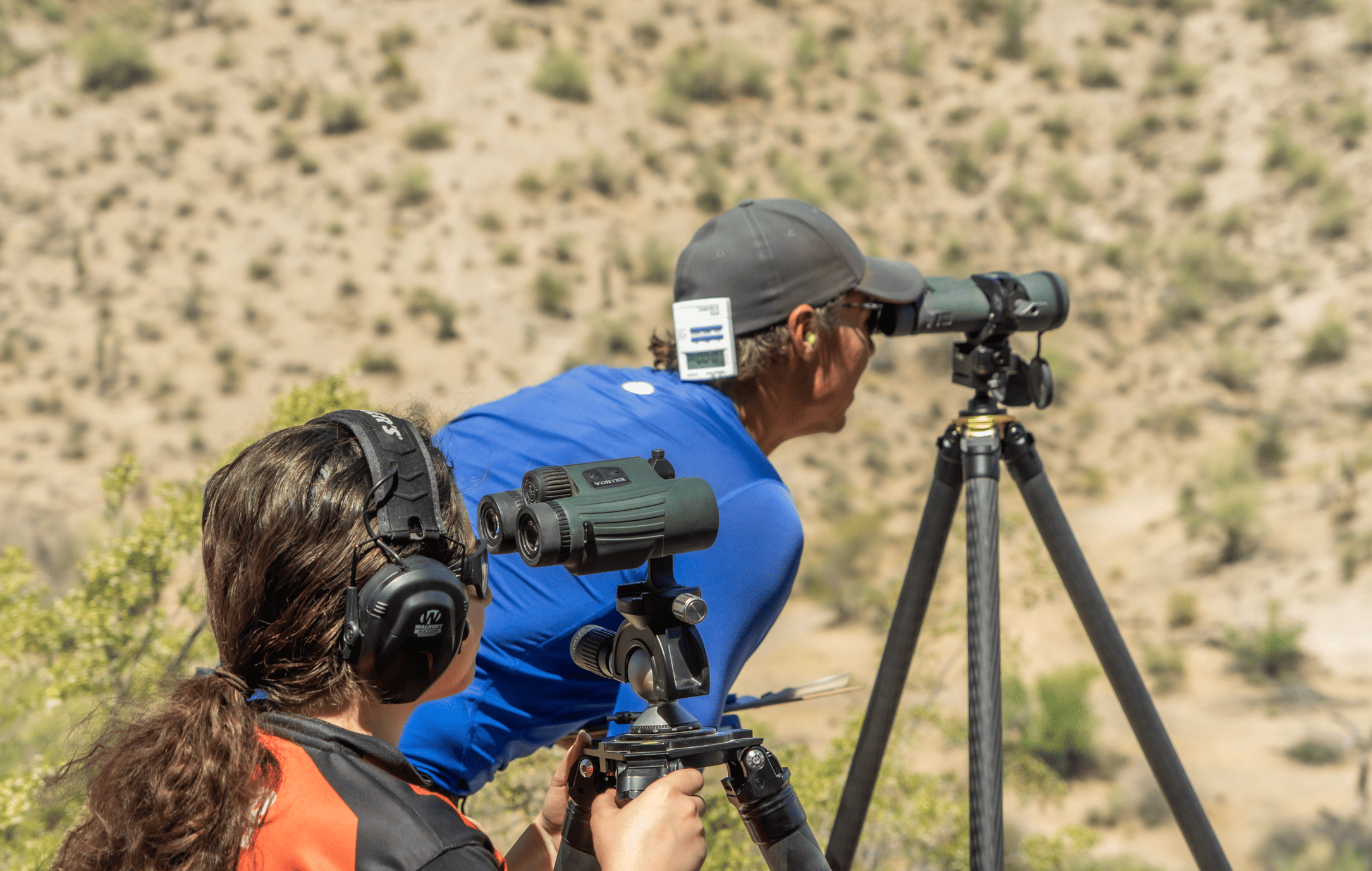 A shooter wearing ear protection uses a tripod-mounted binocular system alongside a spotter utilizing a high-power spotting scope to track impacts downrange.