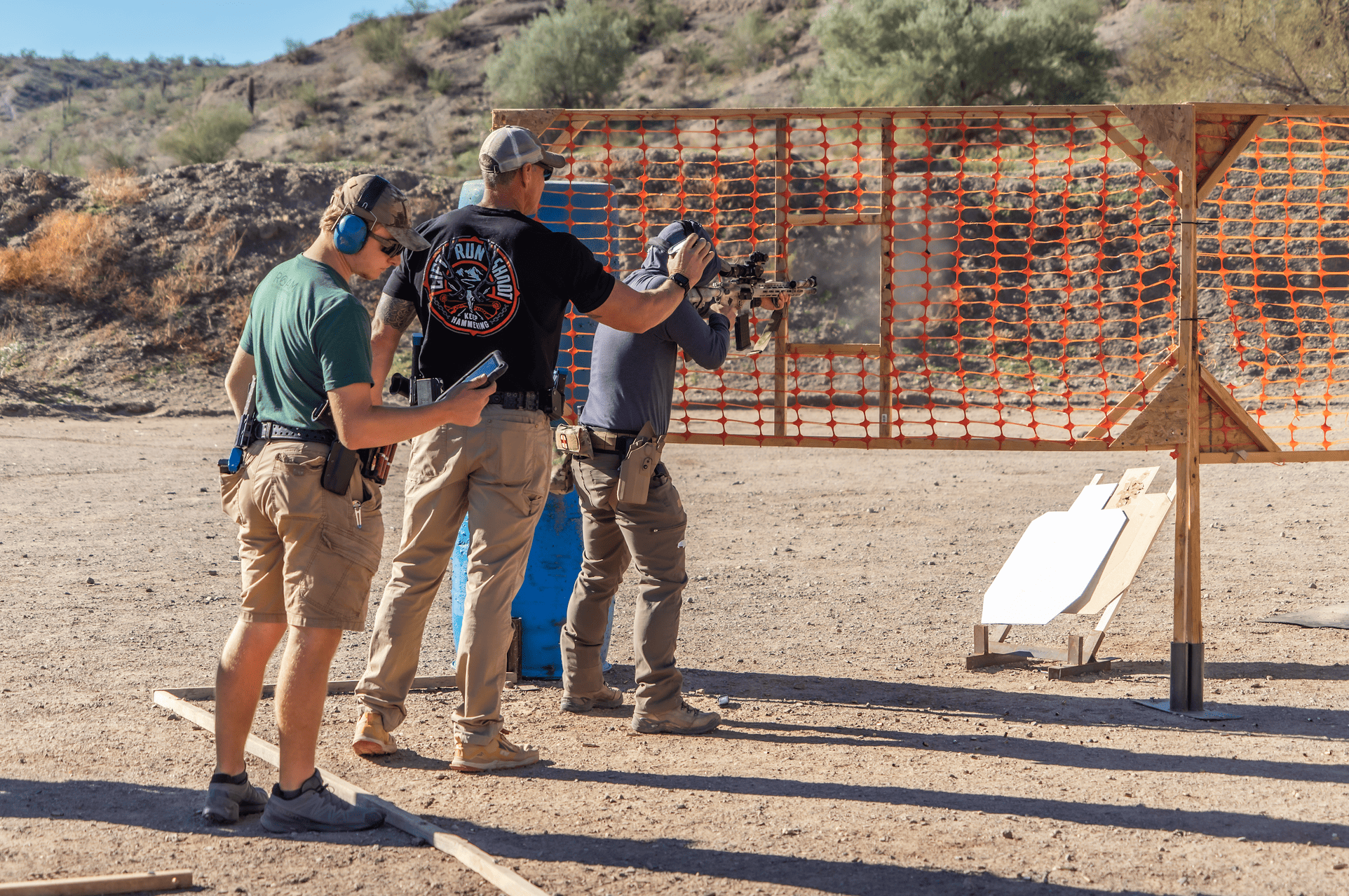 Hands-on training on the range deck featuring a shooter running an AR-platform rifle drill through an orange mesh safety barrier, supported by an instructor in a bold graphic tee and a range officer tracking performance data on a tablet.