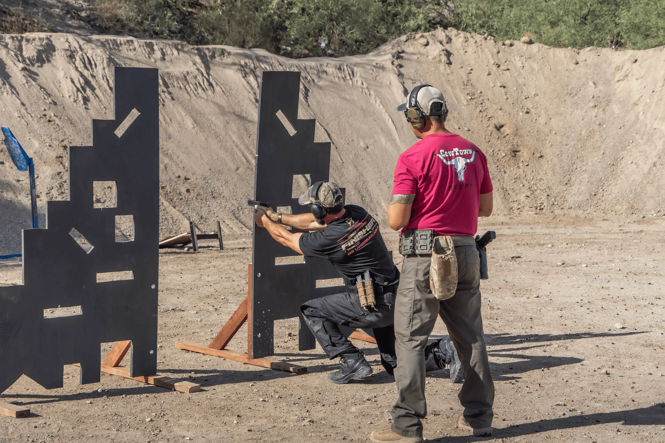A shooter at Cowtown Range practices drawing from a holster and engaging targets through a multi-height wooden barricade, supervised closely by a Cowtown Range Safety Officer wearing a red staff t-shirt on a high-desert tactical bay.