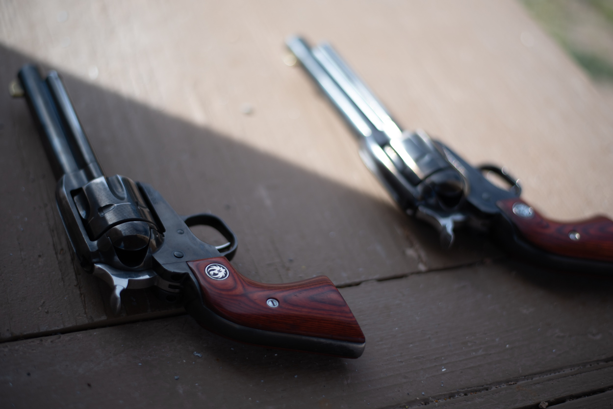 a0b291e2-a18a-4c0e-91de-55b4ab8ad255.webp Two single-action Ruger revolvers rest diagonally on a flat, brown wooden surface, positioned in a mirrored layout. The firearm in the foreground is cast in deep shadow, highlighting its dark blued steel construction and polished, reddish-brown wooden grip panels featuring a silver Ruger phoenix medallion. In contrast, the second revolver lies in the sunlit background, its polished steel barrel and cylinder reflecting the bright light while showcasing identical wooden grips and matching silver logo inlays.