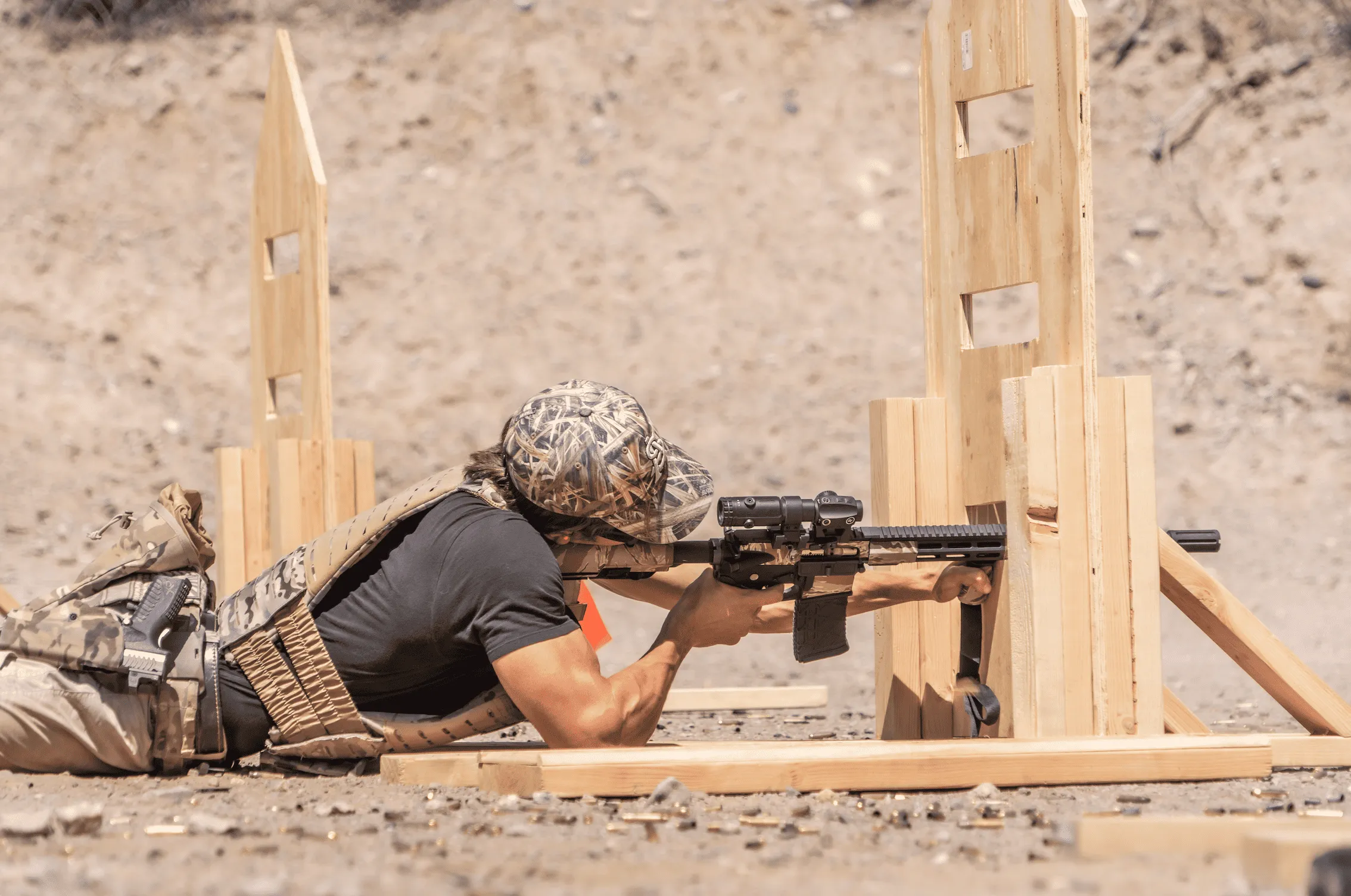 An operator in a prone position wearing a plate carrier and camo cap, actively stabilizing and aiming a camo-patterned carbine platform through a wooden barricade cutout during a dynamic training evolution in an outdoor desert bay.
